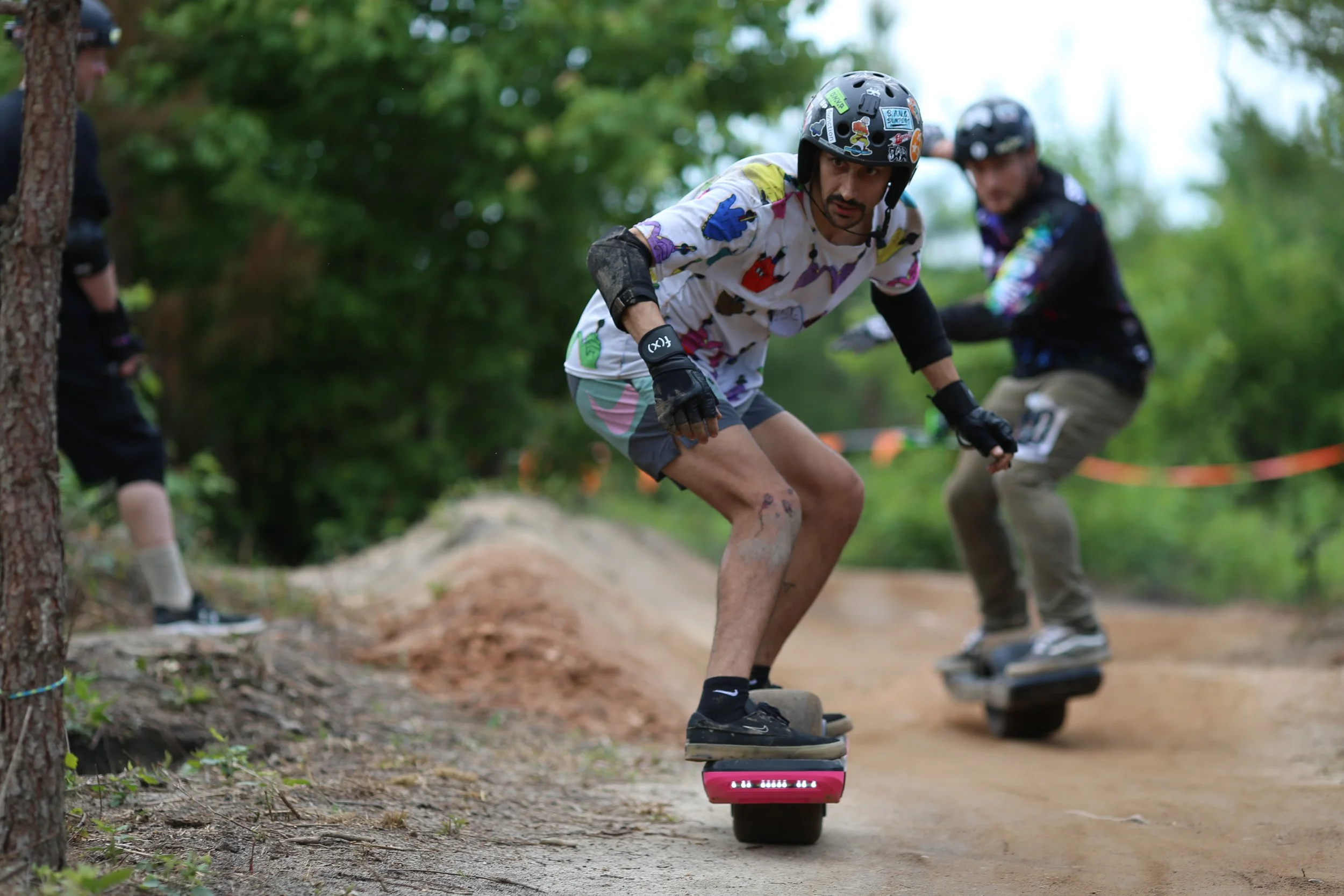 Two people riding electric skateboards on a dirt track, wearing helmets and protective gear, surrounded by trees.