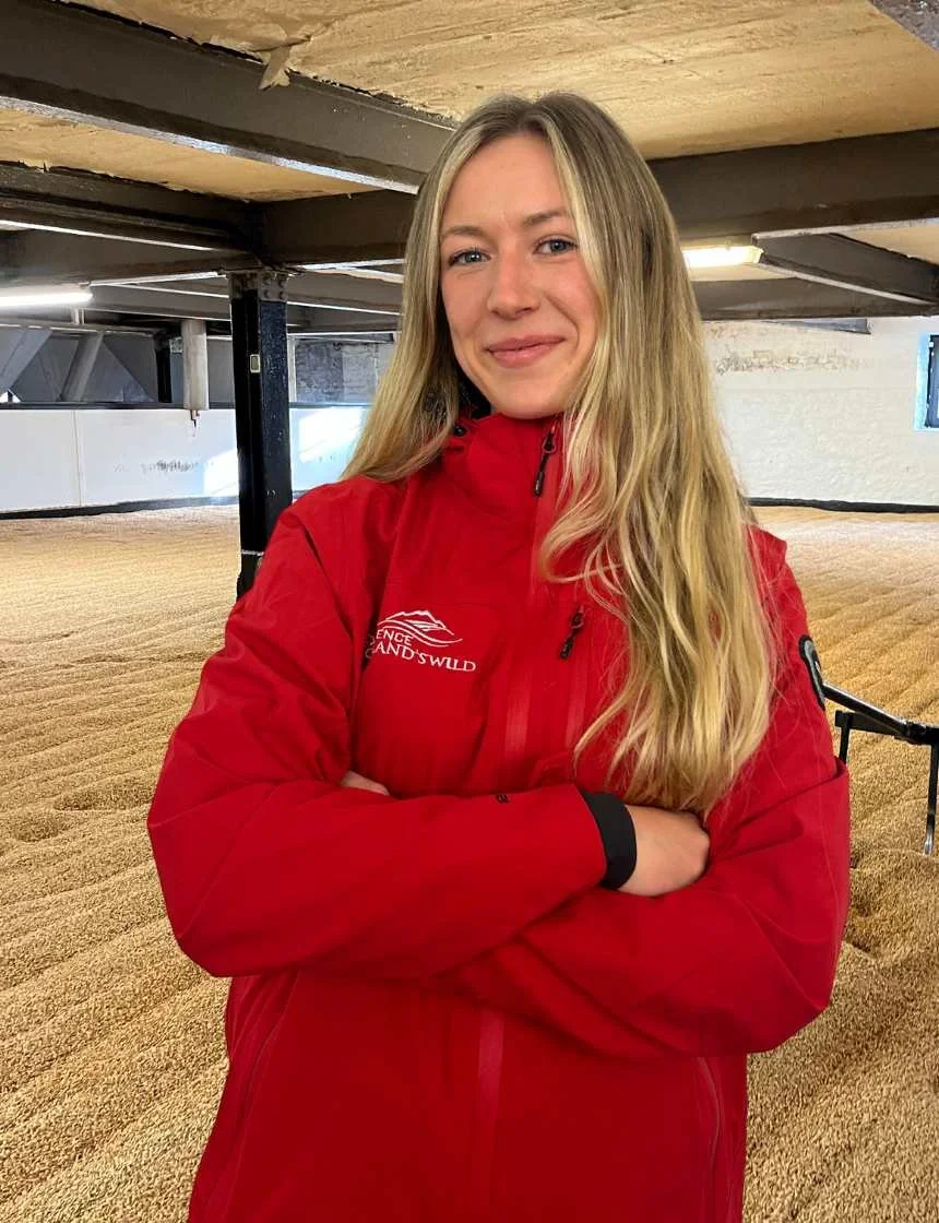 Young woman with long blonde hair wearing a red jacket, standing indoors with her arms crossed, in a setting that appears to be a stable or barn with straw-covered flooring.