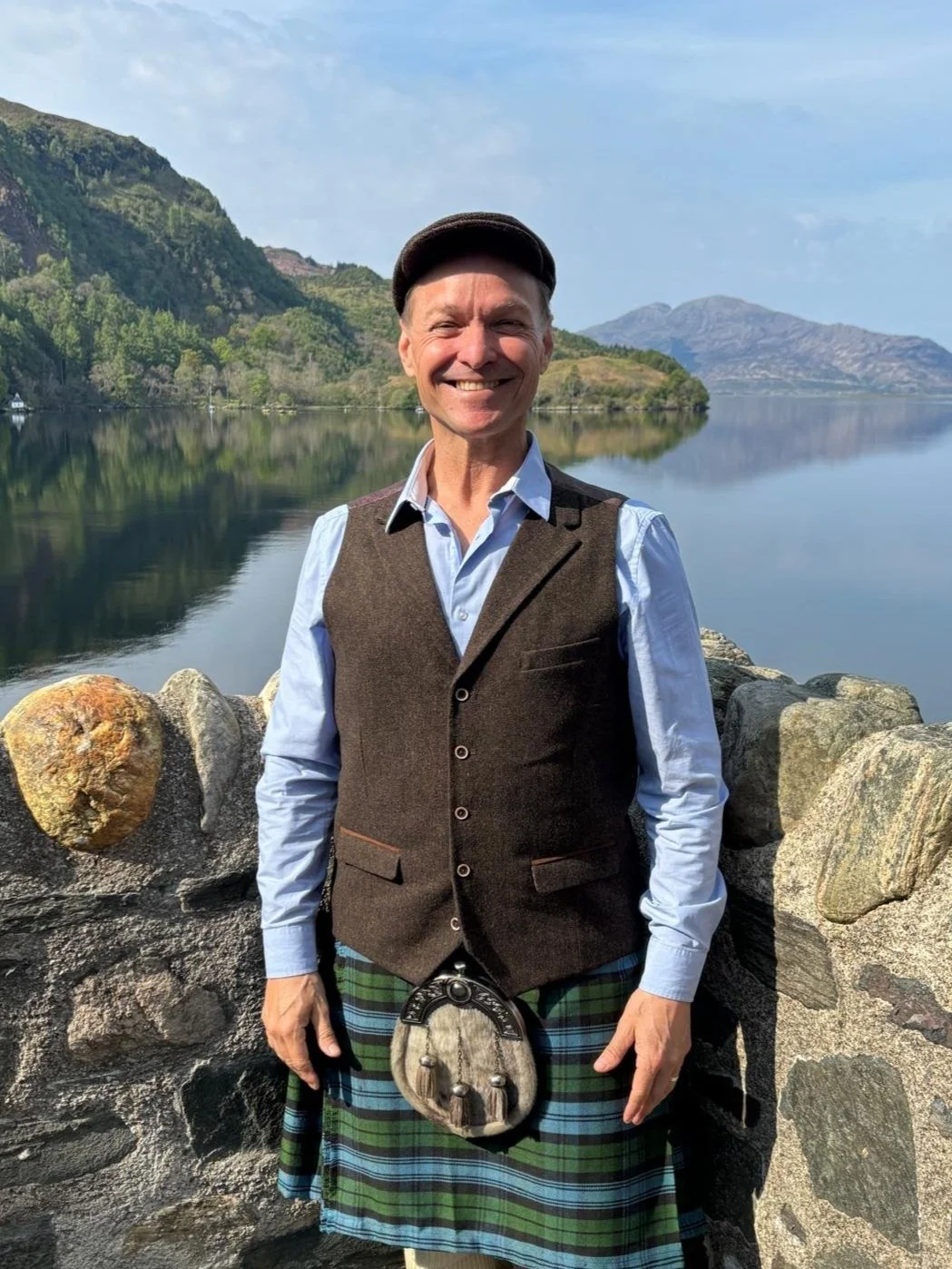 A man dressed in traditional Scottish attire, including a kilt, standing by a lake with mountains in the background.