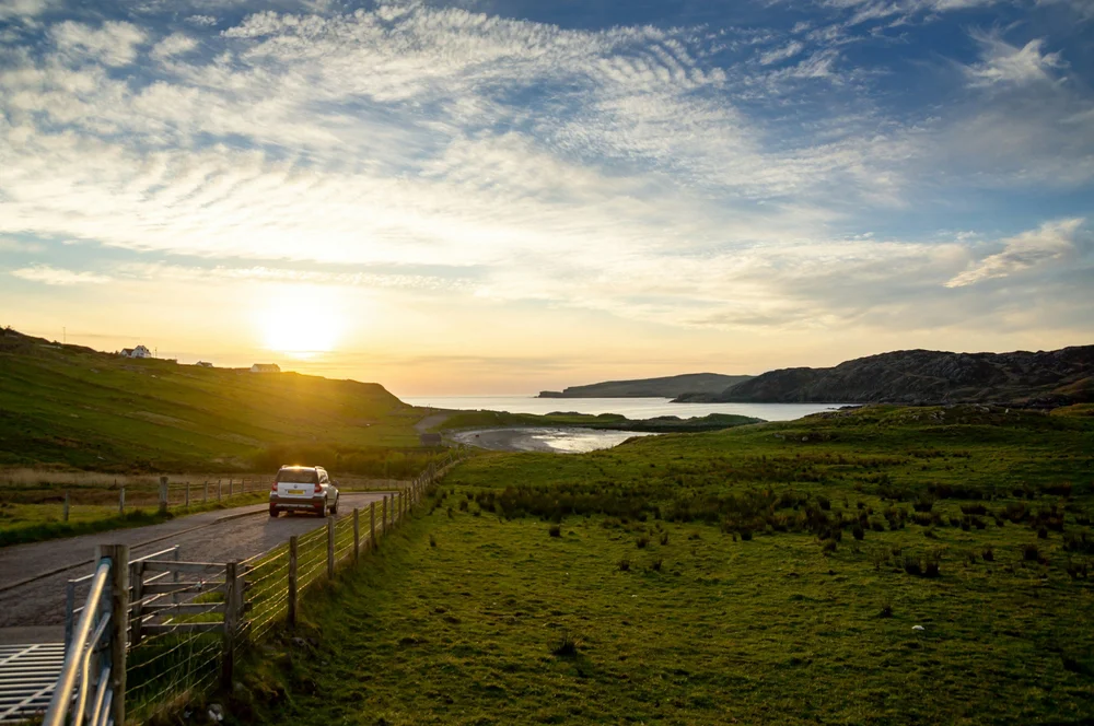 A car on a narrow rural road beside the Scottish coast at sunset