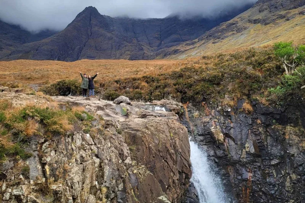 Two travellers waving above a waterfall at the Fairy Pools with the Cuillin mountains behind