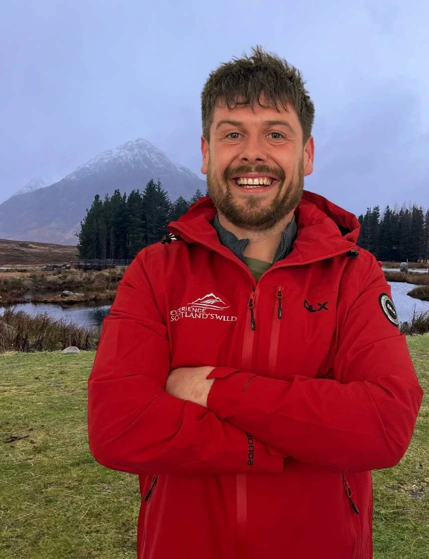 Man smiling wearing a red outdoor jacket with a mountainous landscape in the background.