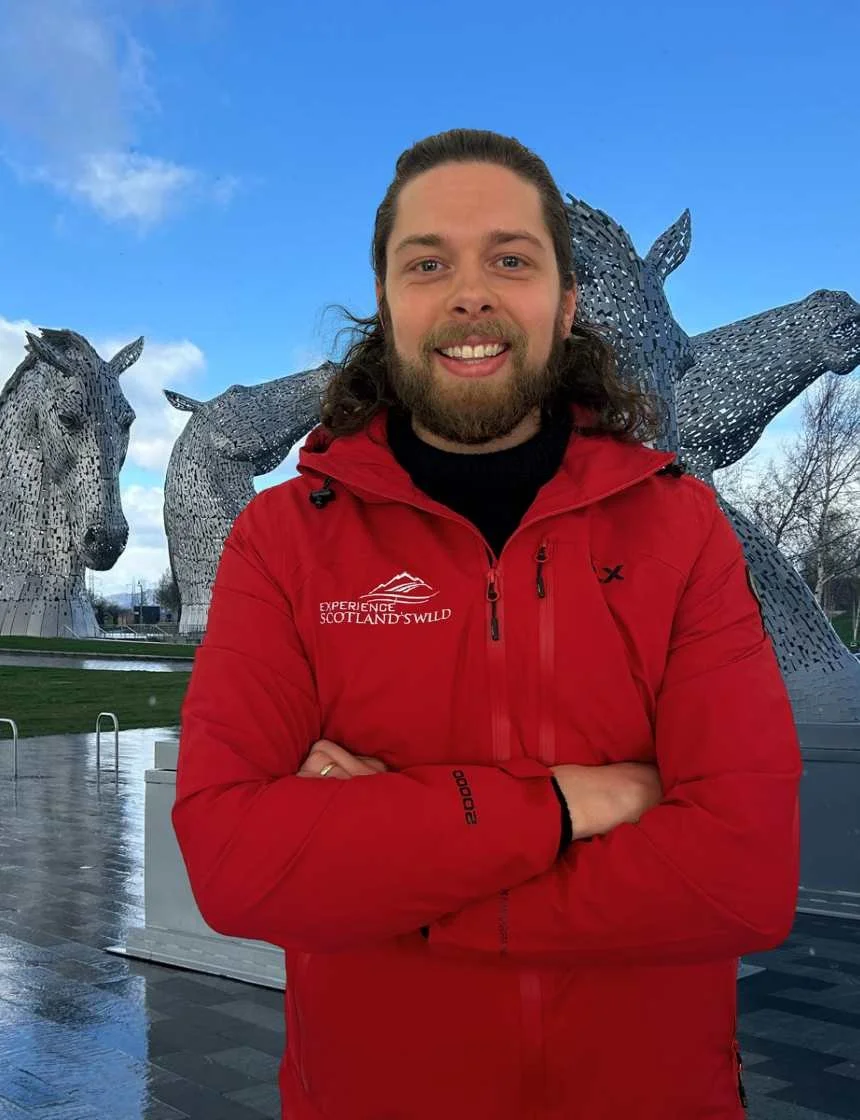 A smiling man with long hair and a beard wearing a red jacket with Scottish wildlife branding, standing with arms crossed in front of the Kelpies, large horse head sculptures in Scotland under a blue sky.