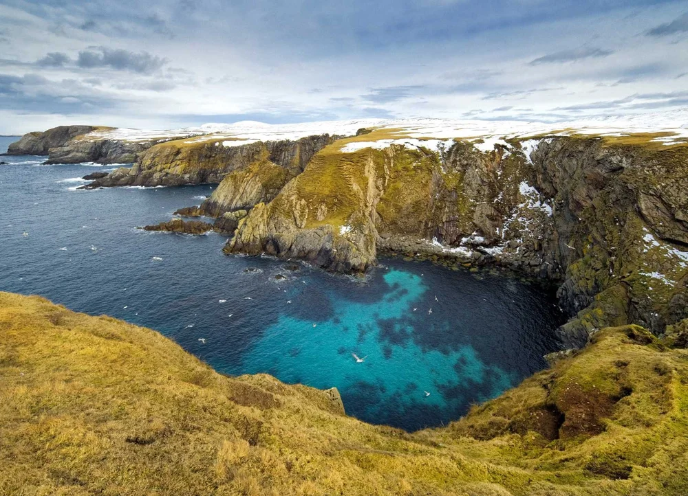 Green-topped sea cliffs around a turquoise inlet on the Shetland coast