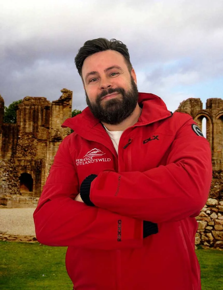 A man with a beard and dark hair, wearing a red jacket with patches and text, standing outdoors in front of ancient ruins, with arms crossed and a slight smile.