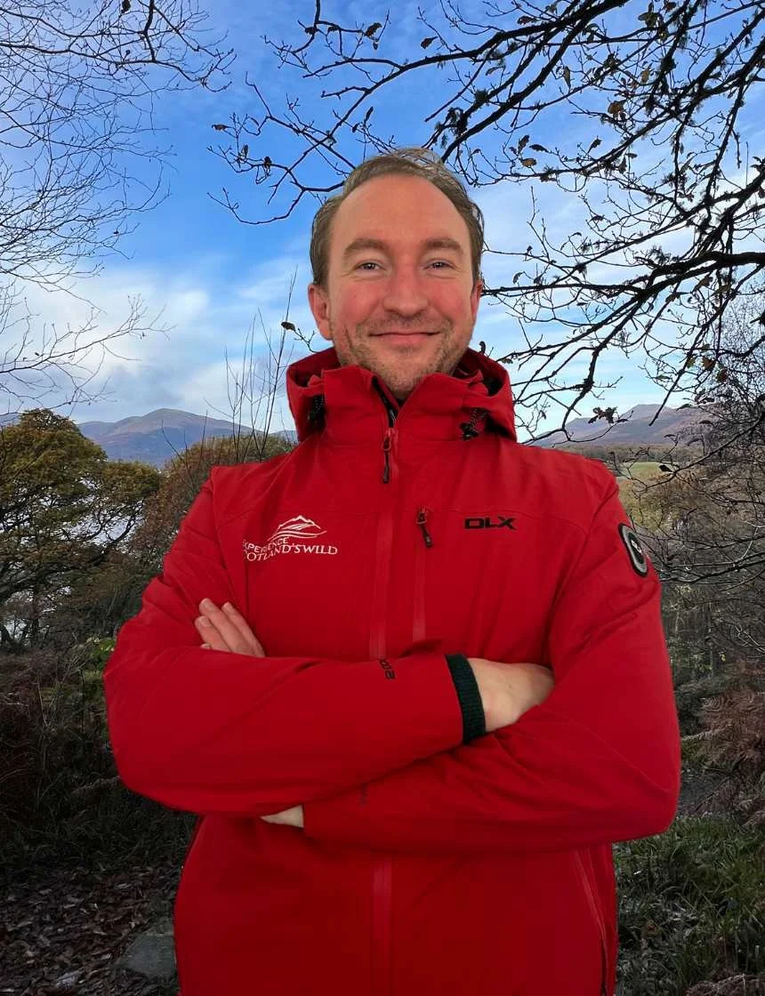 Smiling man outdoors on a mountain trail during autumn, wearing a red outdoor jacket, with leafless trees and mountains in the background.