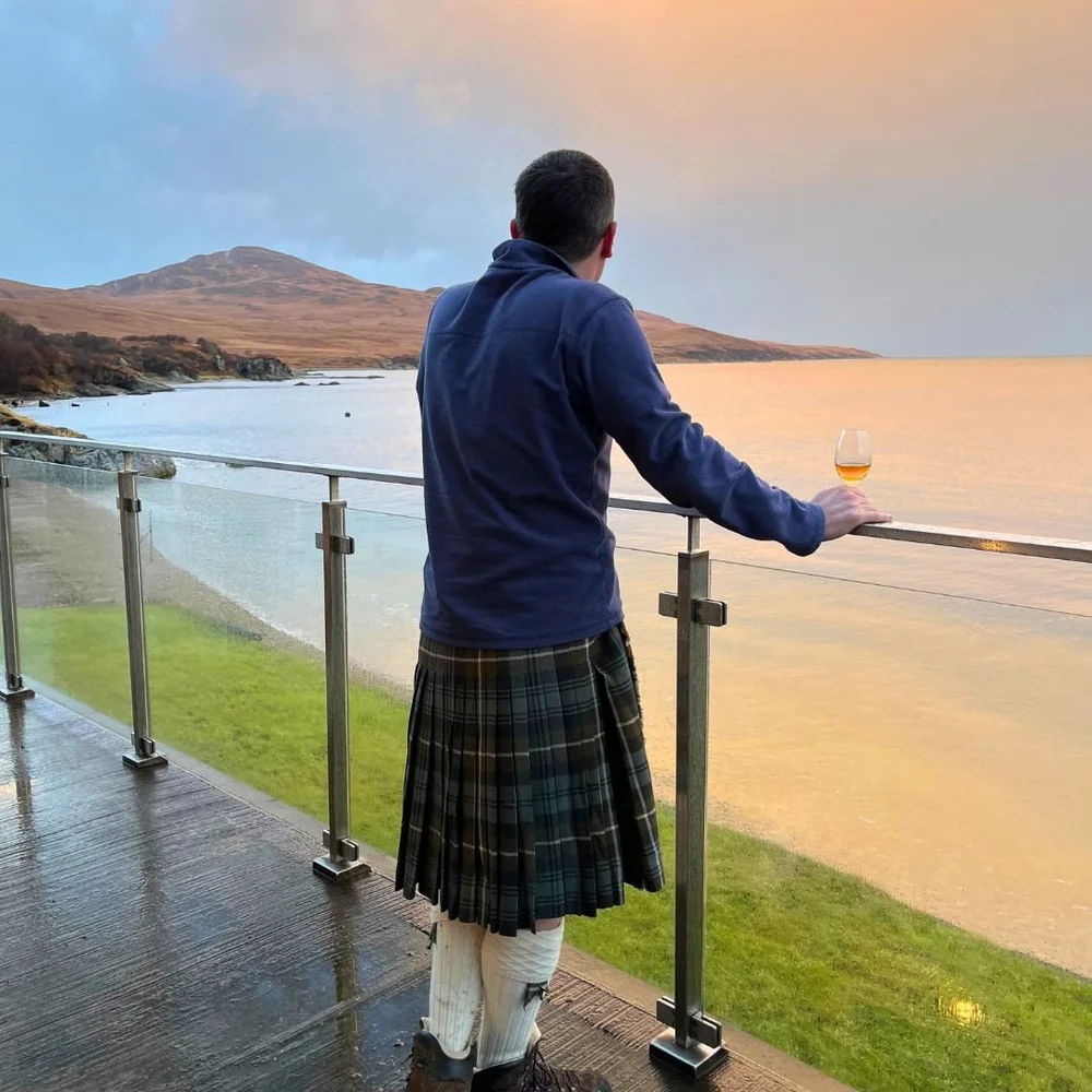 A kilted visitor with a whisky glass looking across the water on Islay