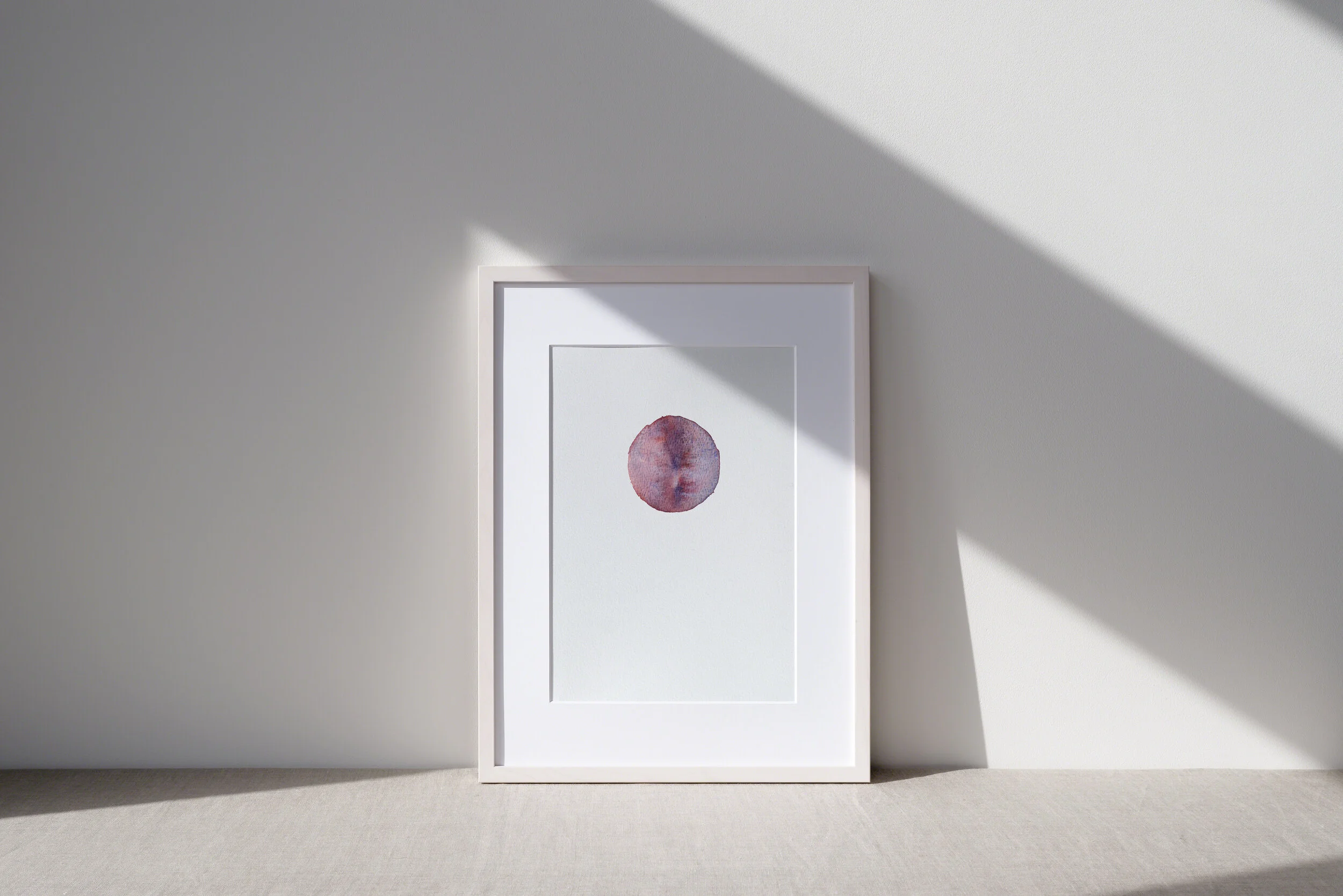 Framed artwork of a watercolor-style circle with shades of purple and pink, leaning against a wall with sunlight casting shadows.