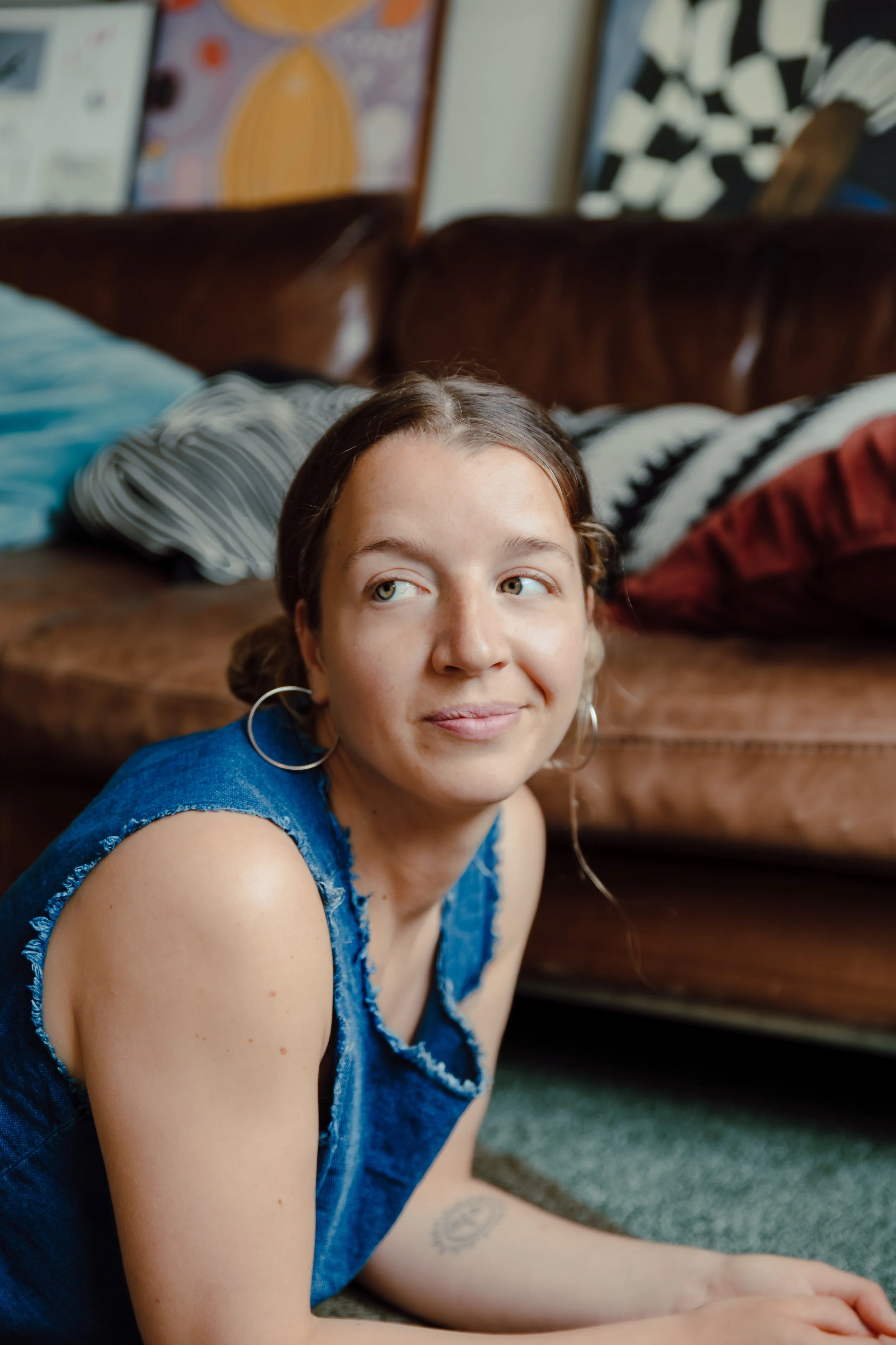 Young woman with earrings and a blue sleeveless top, lying on her stomach on the floor in a living room, smiling while looking off to the side, with a brown couch and artwork in the background.