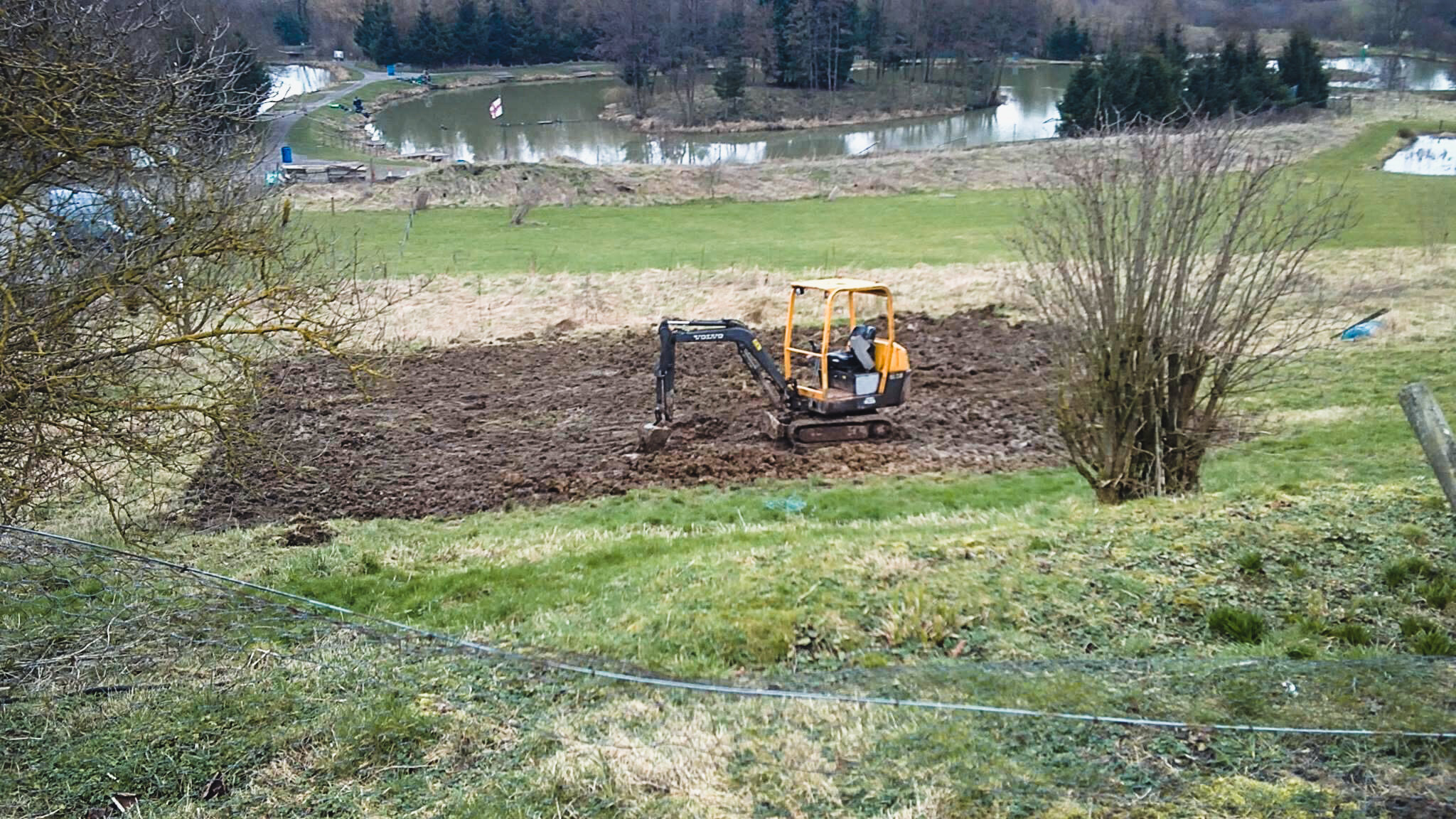 Small orange and black excavator working on a patch of dirt in a grassy area surrounded by trees, with a river and more trees in the background.