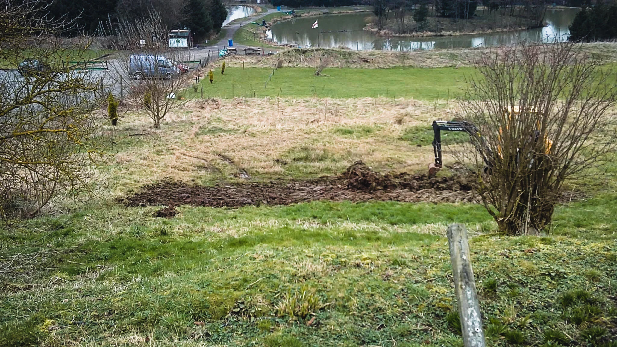 A grassy landscape with small trees, a construction excavator working on a trench, and a pond in the background.
