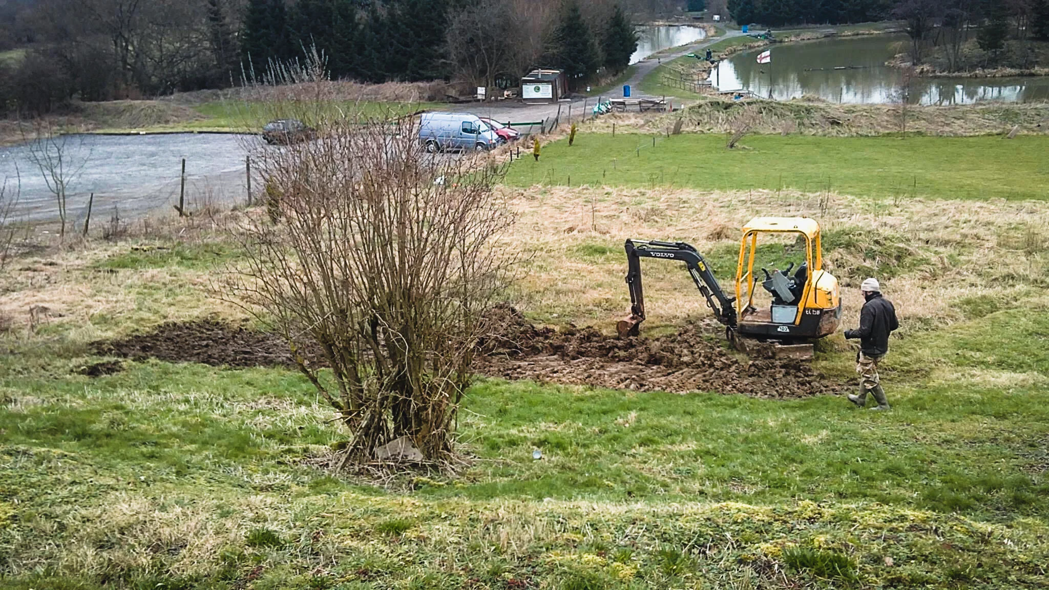 Person operating a small bulldozer on a grassy hillside near a park and a pond.