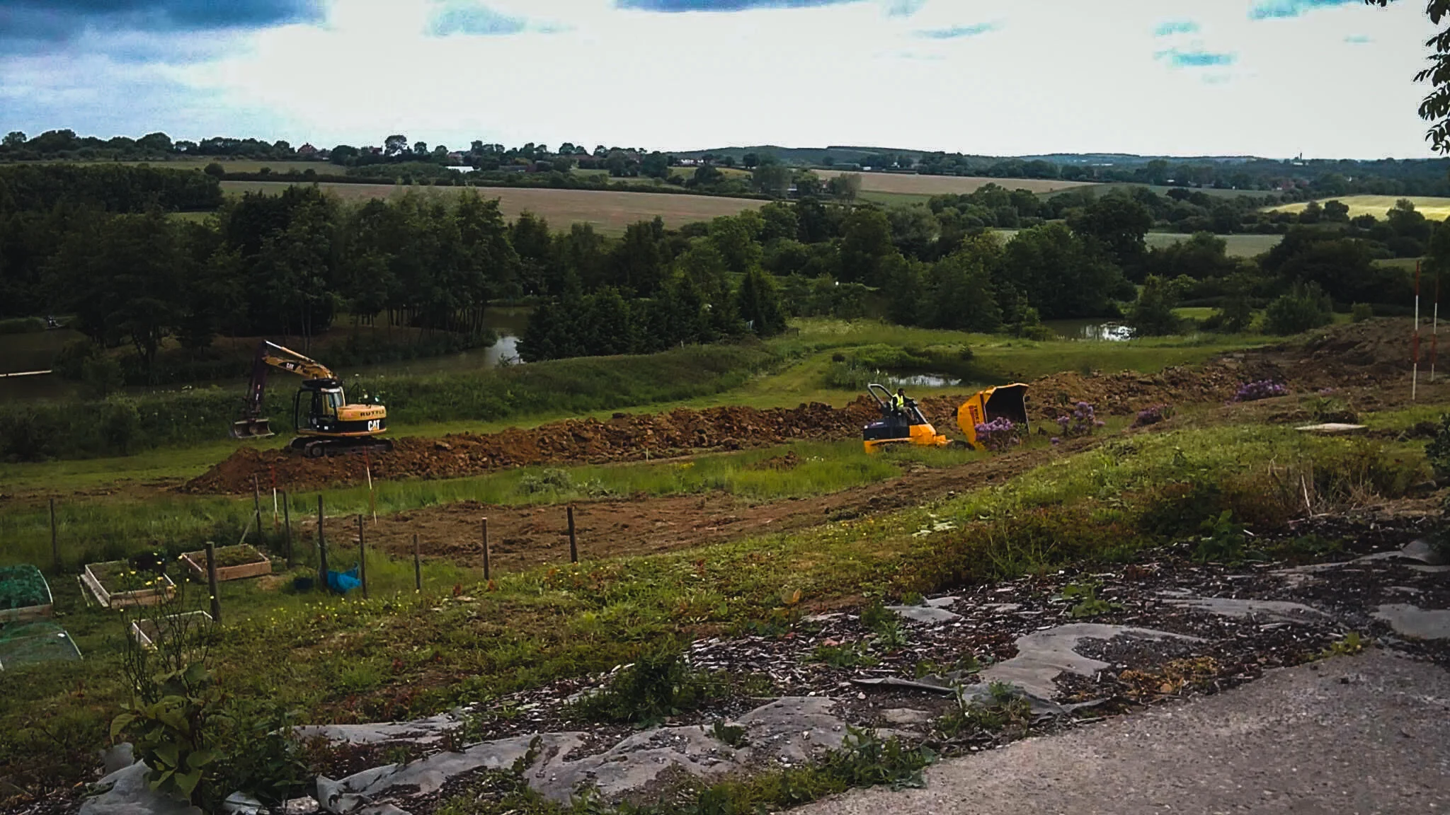 A construction site in a rural area with excavator and mini bulldozer working on dirt, surrounded by greenery, trees, and a pond in the background.