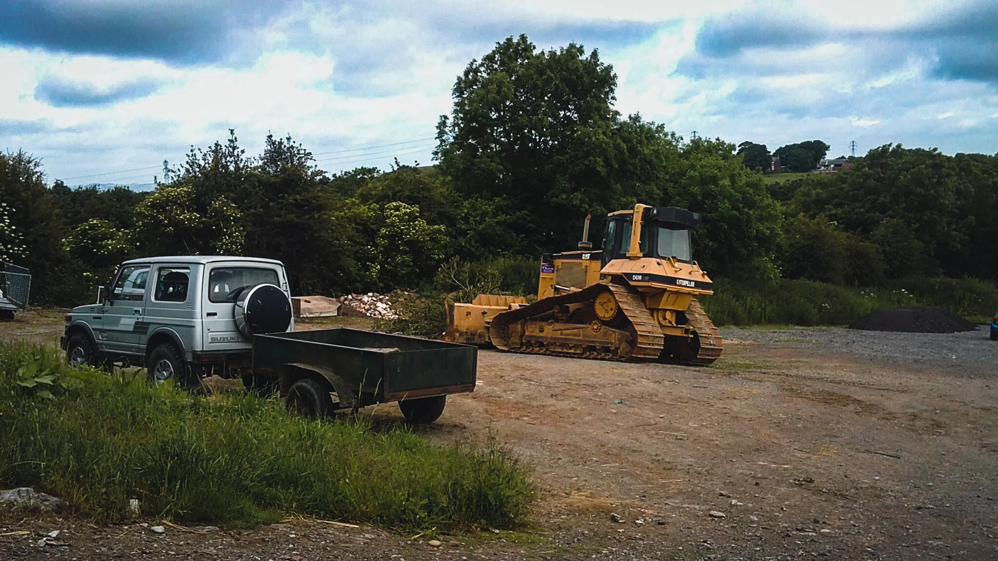 Construction site with a yellow bulldozer, a silver utility vehicle, and a green trailer on dirt ground, with trees and cloudy sky in the background.