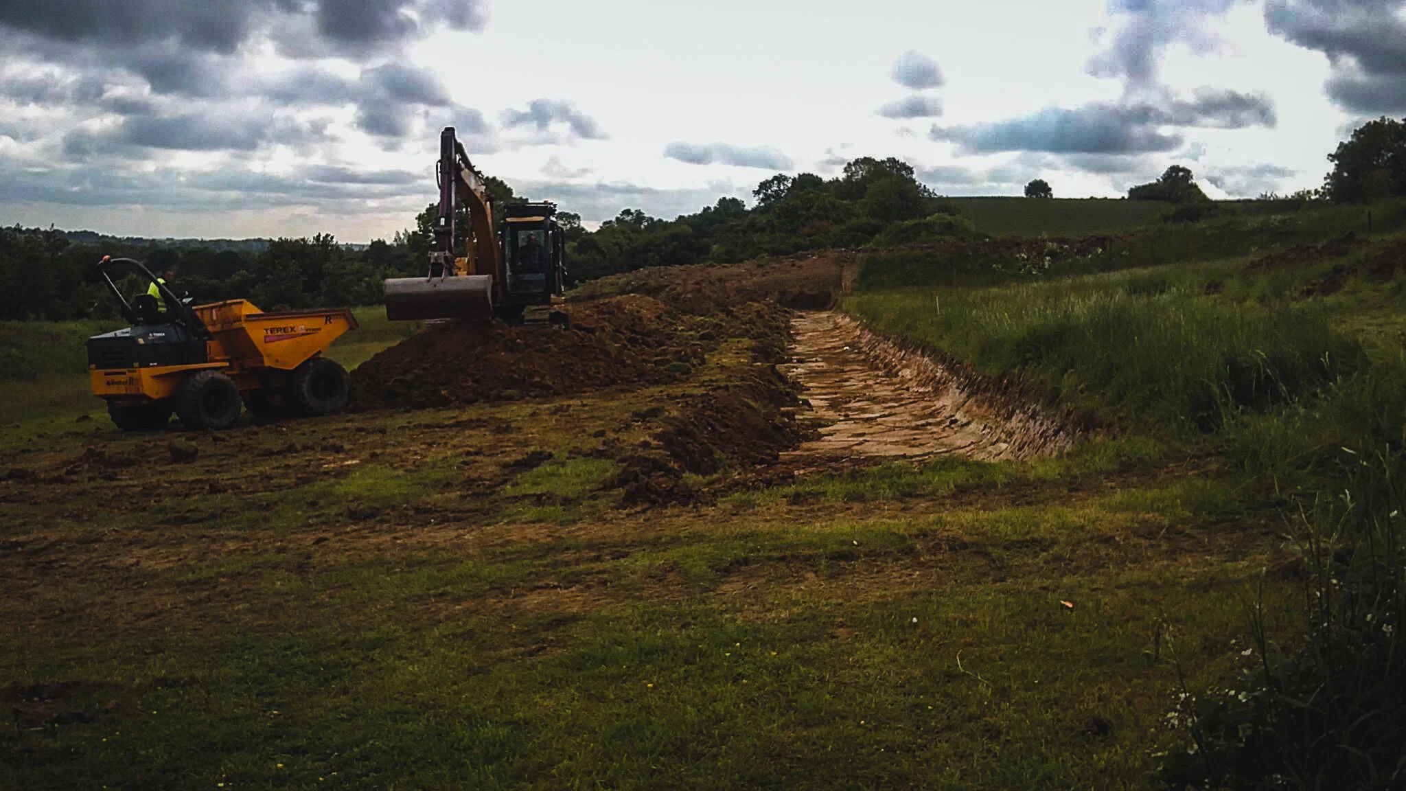 Construction site with excavator and compactor working on a dirt path in a grassy, rural area under a cloudy sky.