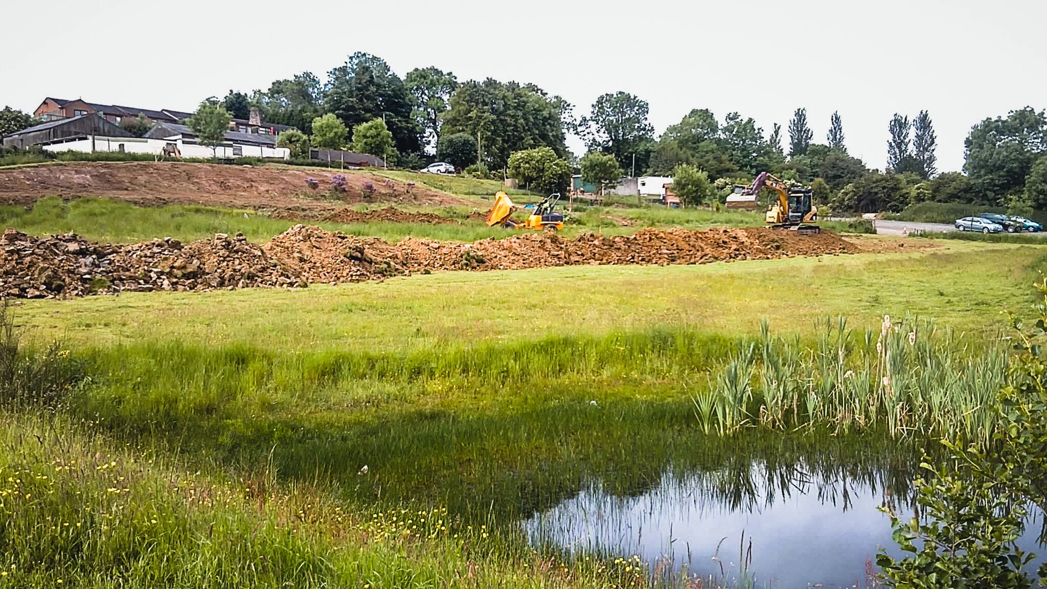 Construction site on grassy field with bulldozer and excavator, dirt piles, houses on hill, and a small pond in foreground.