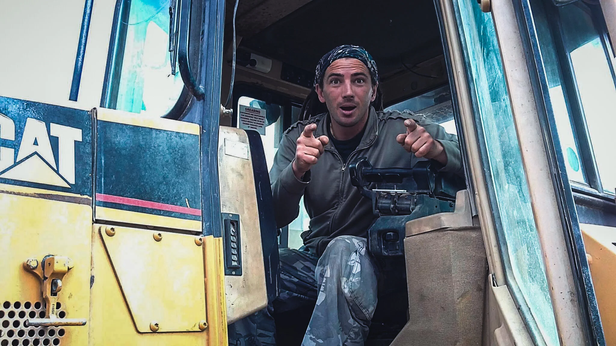A man sitting inside a yellow construction vehicle, pointing and looking surprised or excited.