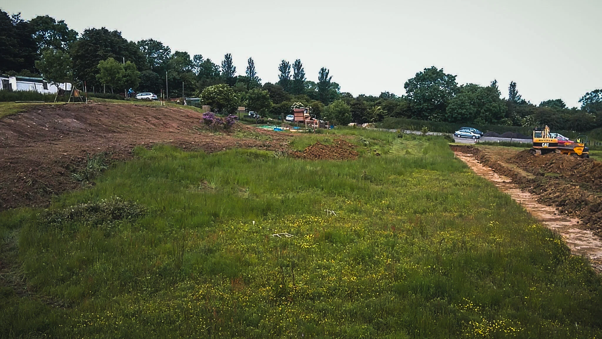 A construction site with a grassy area, a dirt path, and construction equipment, including a yellow excavator, with trees and parked cars in the background.
