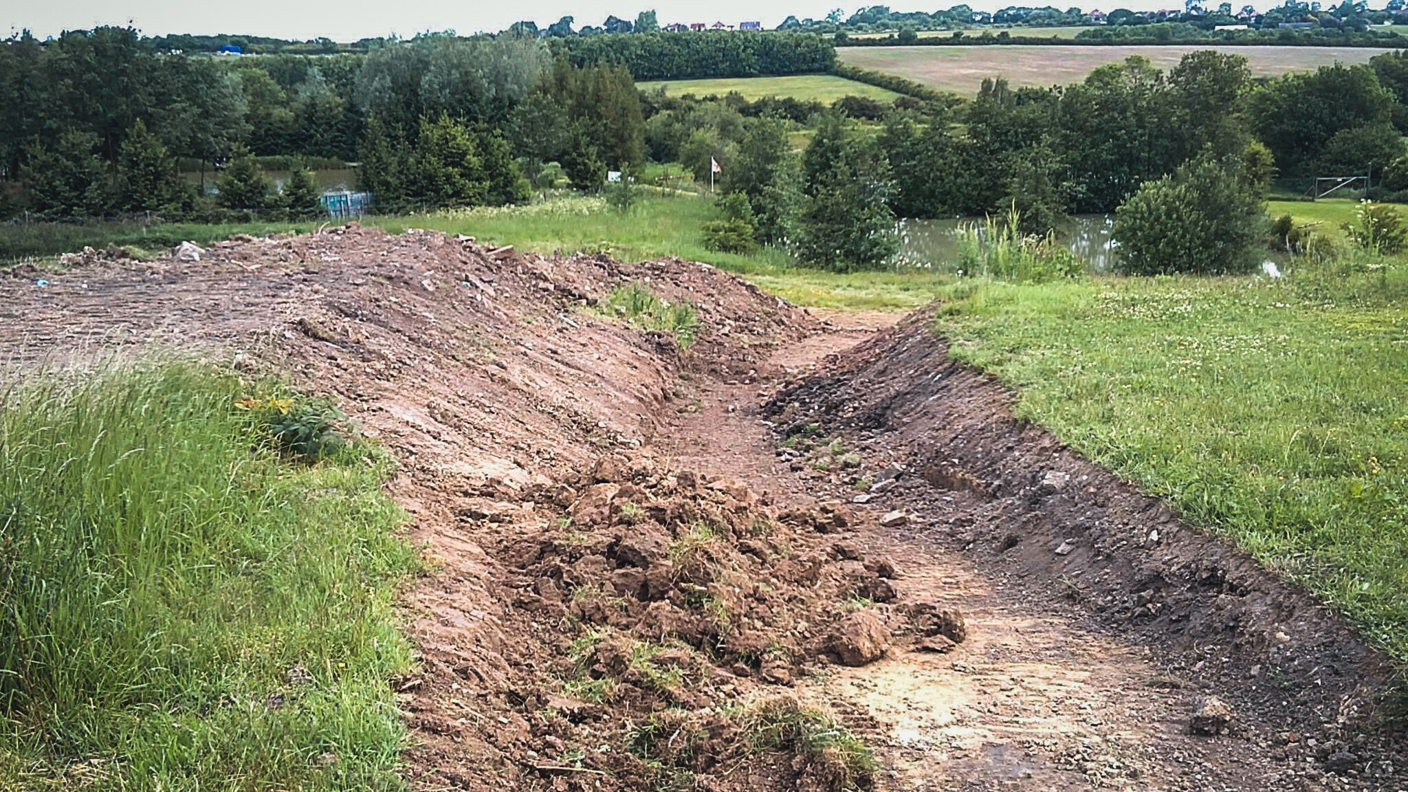 A dirt path or trench in a grassy area near a pond, with trees and fields in the background.