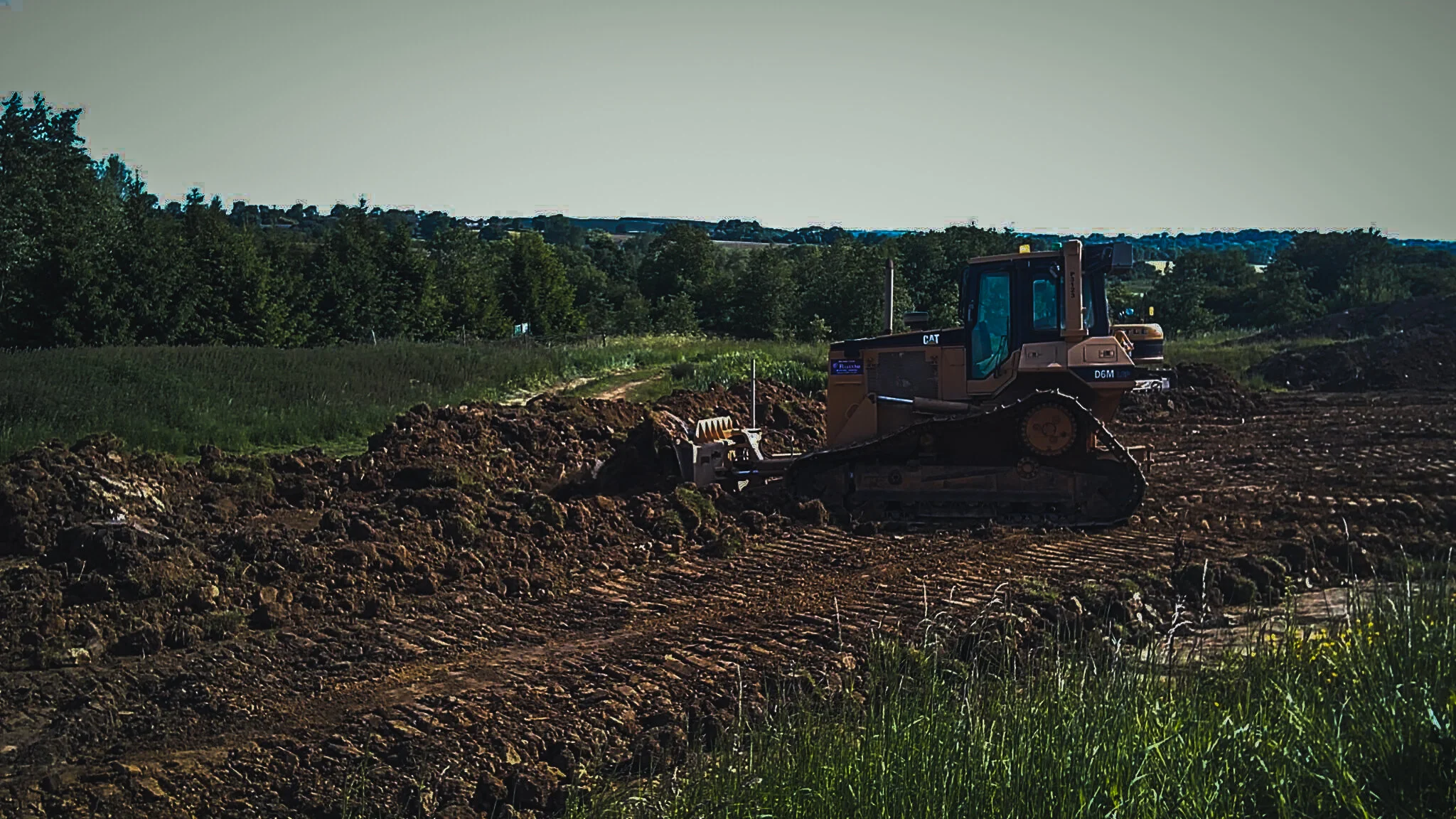 A bulldozer working on a construction site in a rural area with fields and trees in the background.