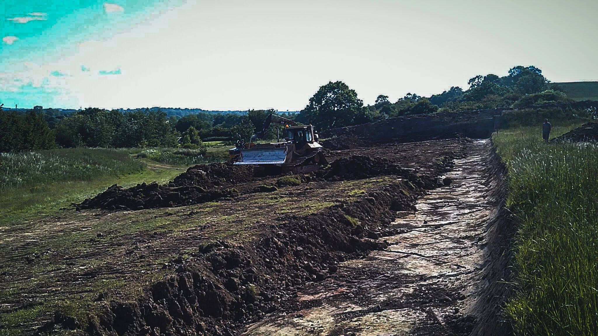 A bulldozer working on a muddy construction site with trees and hills in the background.