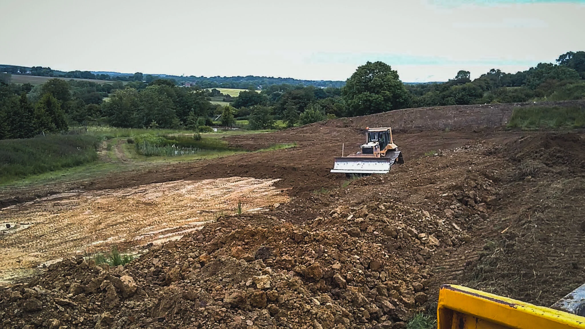 Construction site with bulldozer clearing land, surrounded by green trees and rolling hills in the background.