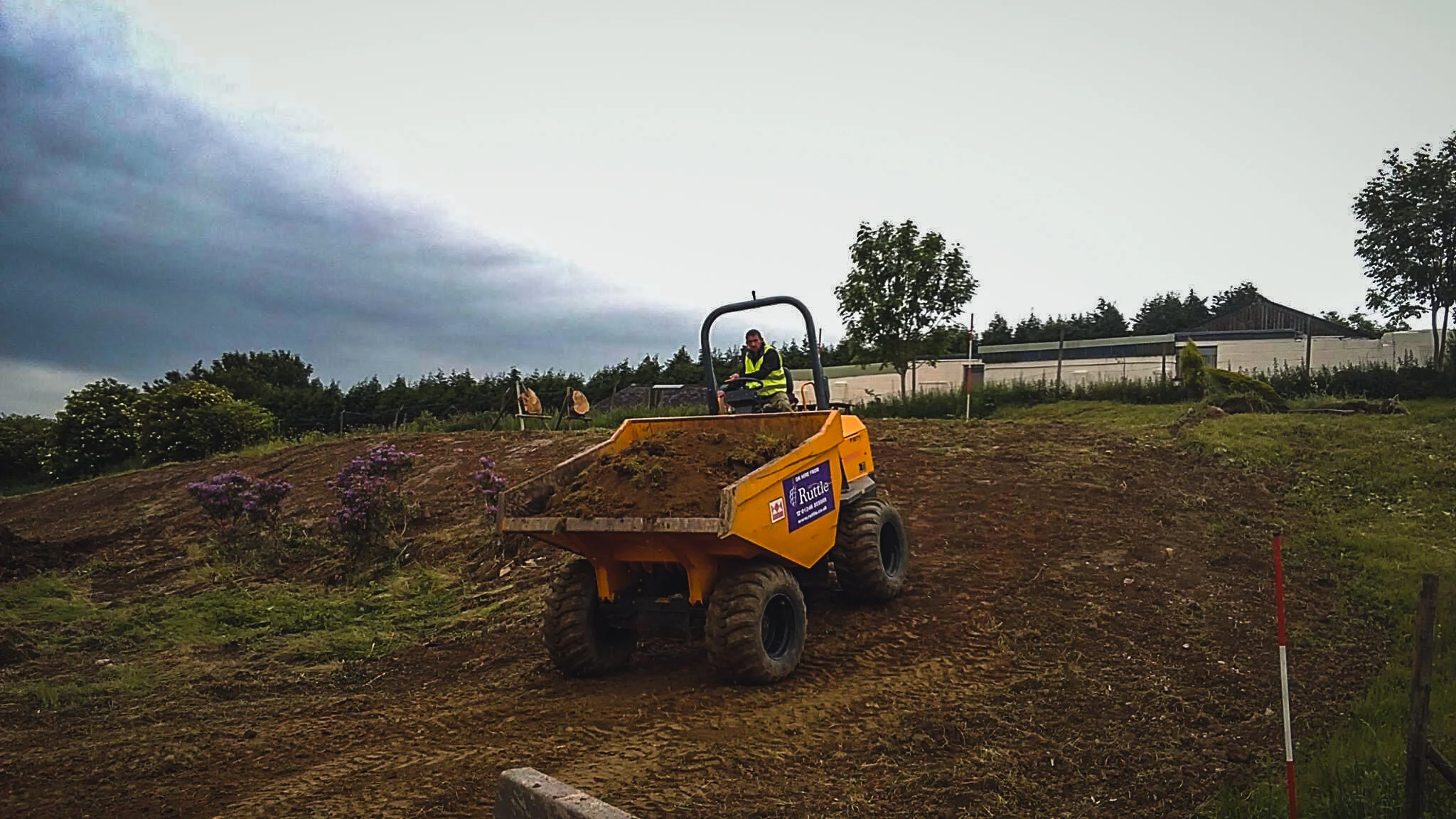 A person operating a yellow dumper truck on a hillside with patches of purple flowers, trees, and a building in the background under a cloudy sky.