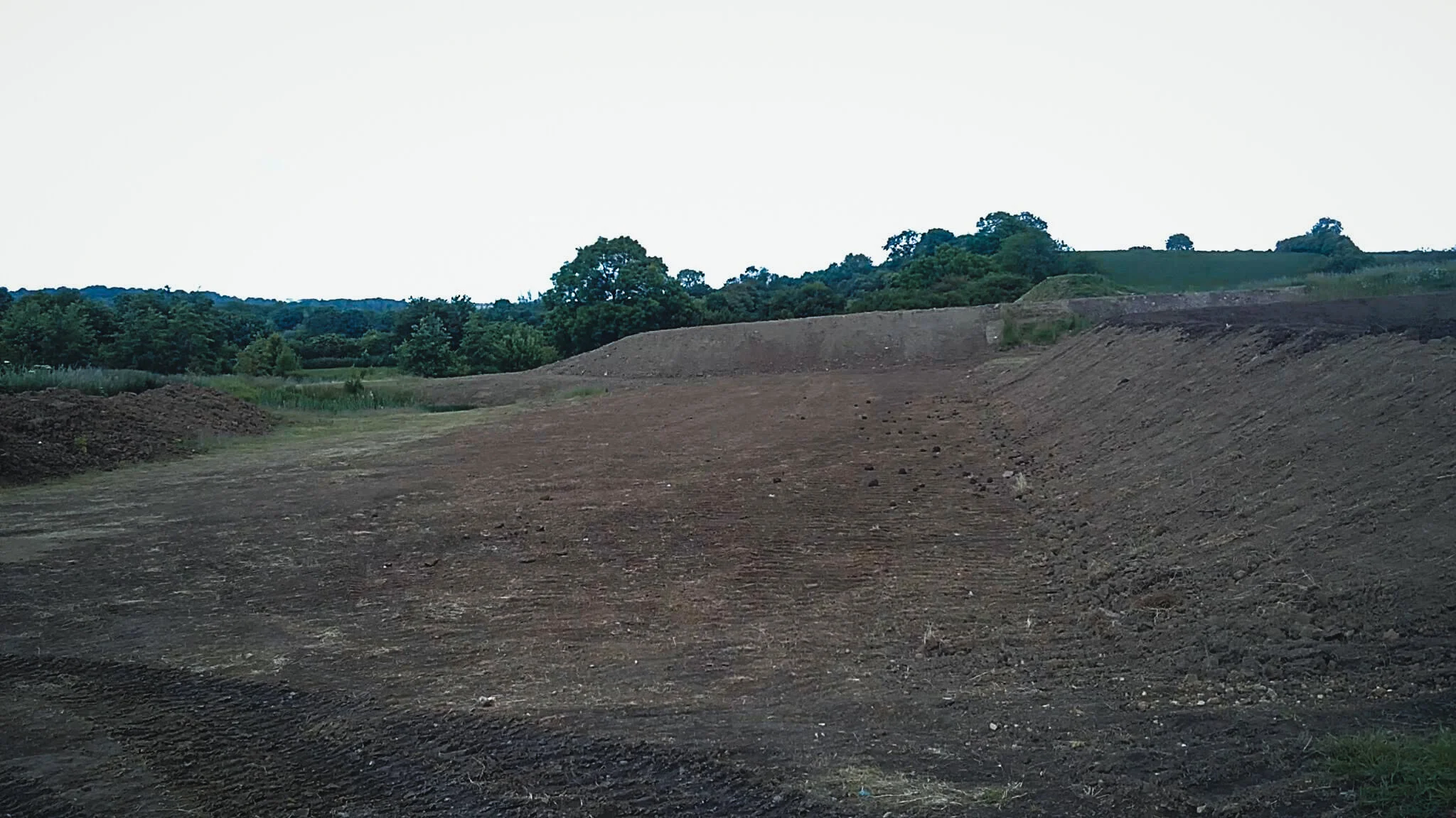 A barren, excavated landscape with mounds of dirt and a backdrop of green trees and shrubs under an overcast sky.