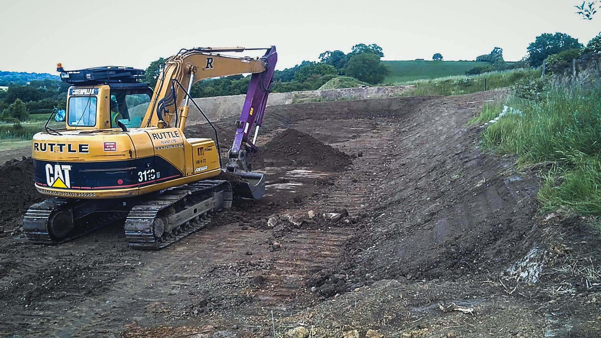 A yellow Caterpillar excavator working on a dirt construction site in a rural area with green fields and trees in the background.