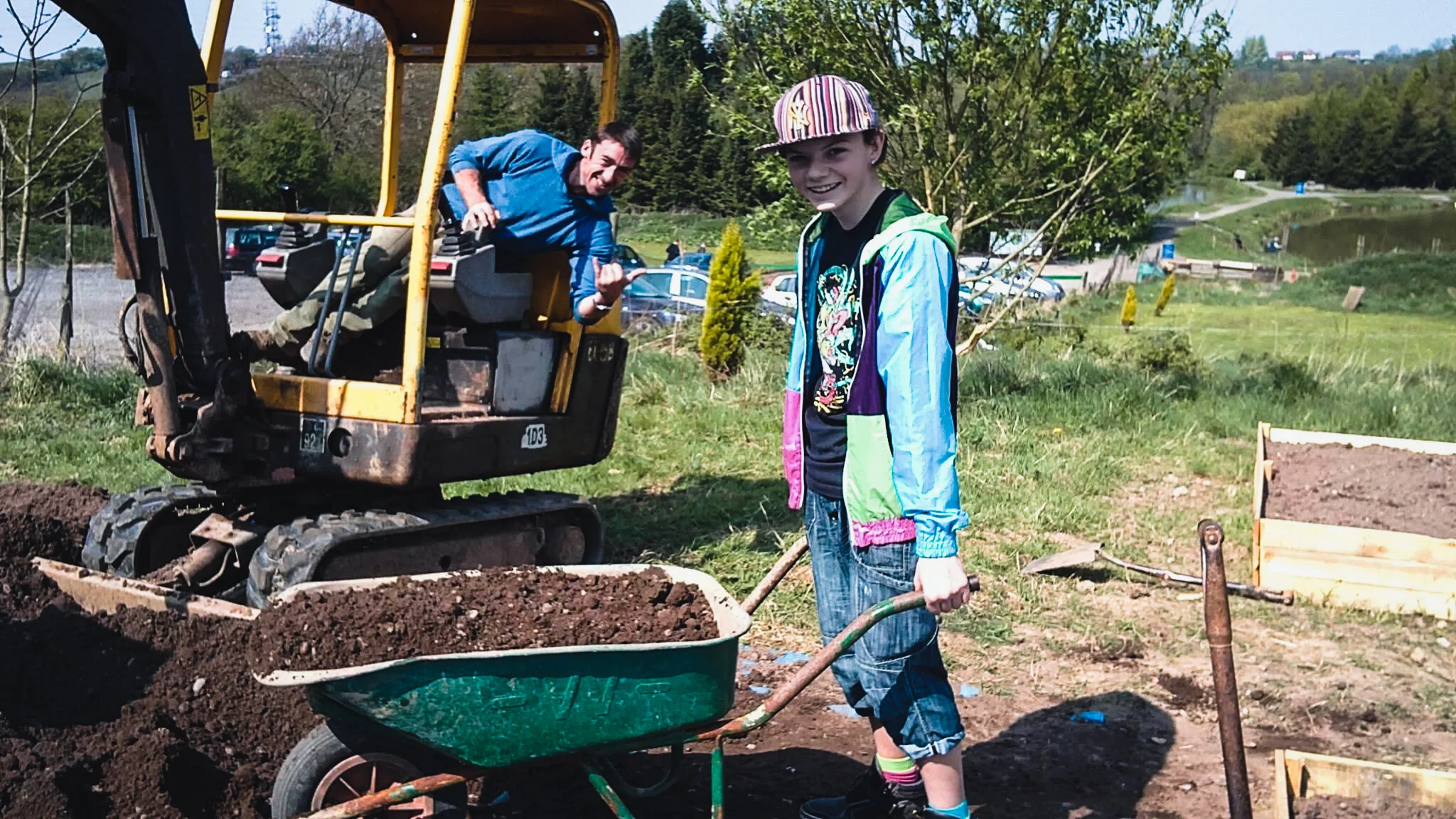 A boy with a cap and a girl in colorful jacket working on a gardening project outdoors, with a small excavator and wheelbarrow filled with soil nearby.