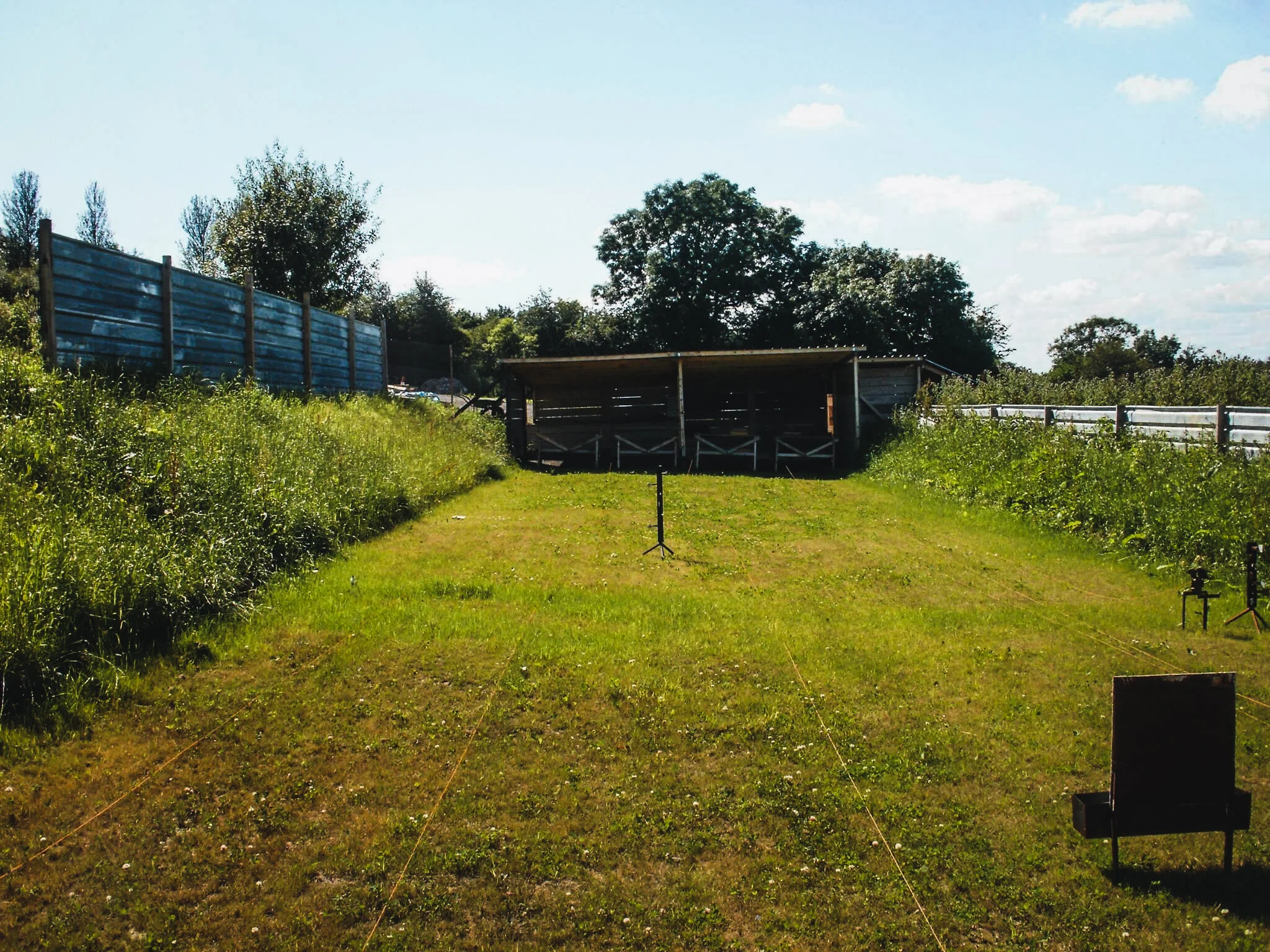 A grassy outdoor shooting range with a covered wooden target area at the end of the range, surrounded by a fence and lush greenery under a partly cloudy sky.