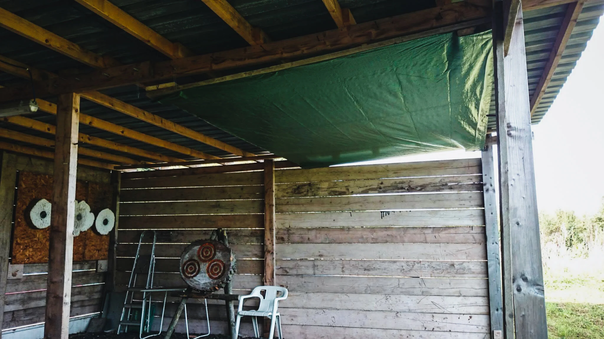 Wooden outdoor structure with a green tarp across the ceiling, a target on a chopping block, a white plastic chair, and a metal stand in a grassy area.