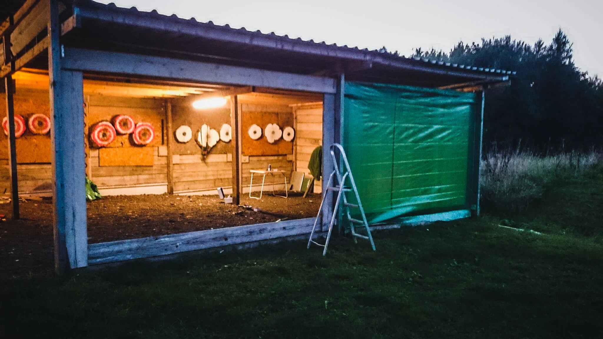 A small wooden shooting range with targets on the back wall, illuminated by a yellow light, and a green tarp covering part of the entrance, with a ladder leaning on the side, surrounding grassy area and trees in the background.