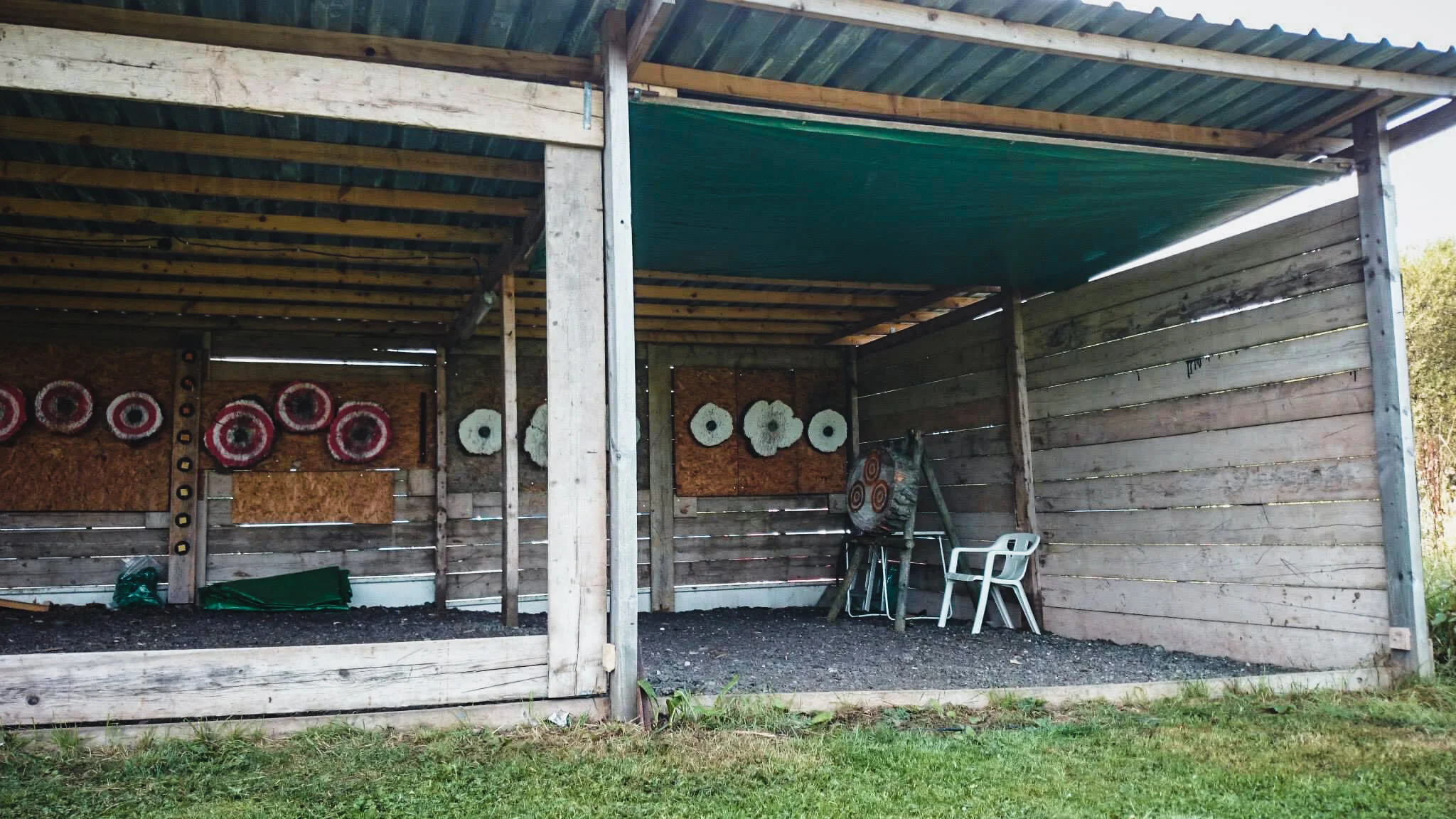Outdoor wooden archery target shed with target boards and archery targets inside, with chairs and greenery outside.