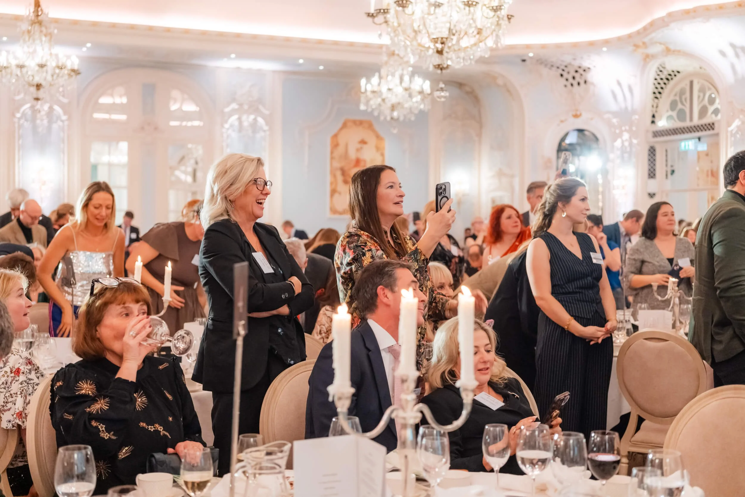 People standing and sitting at a formal event in a grand, ornate ballroom with chandeliers. Some are smiling, taking photos, or drinking, dressed in elegant attire.