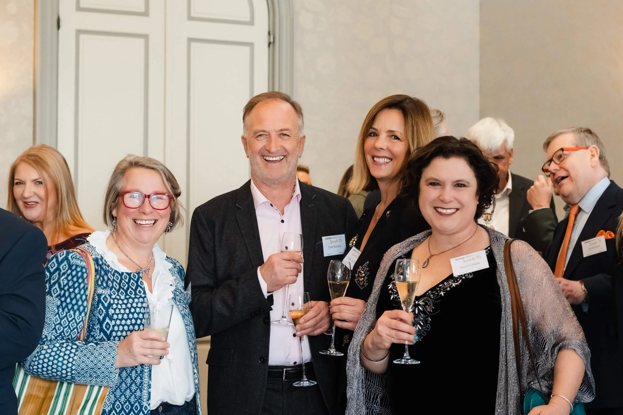 A group of people at a social event, smiling and holding glasses of champagne, dressed in business or semi-formal attire.
