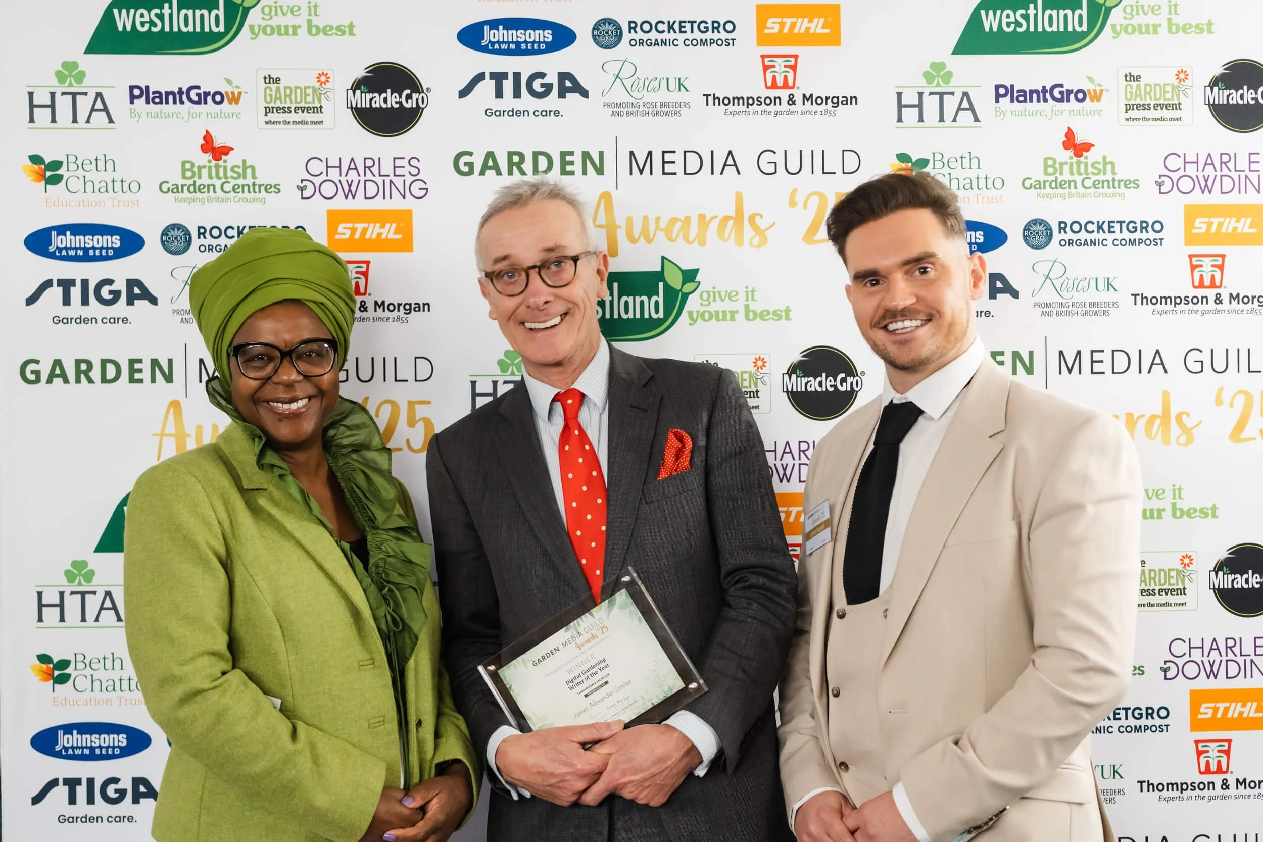 Three people at an award event, two men and one woman. The woman on the left is wearing a green headscarf and matching green coat. The man in the middle is wearing glasses, a dark suit with a red polka-dot tie, and is holding an award plaque. The man