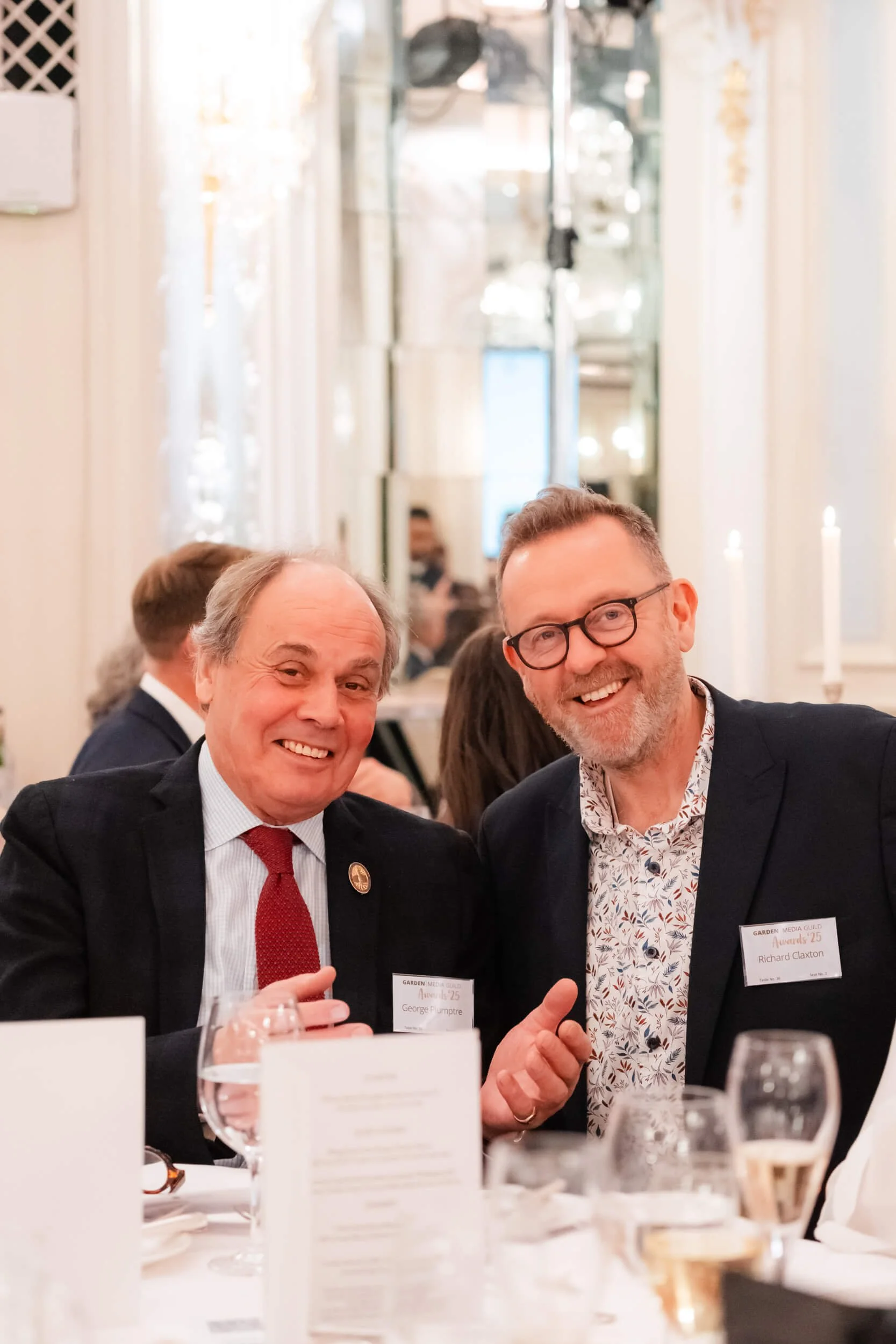 Two men dressed in suits smiling and sitting at a table during a formal event or dinner, with glasses and a menu on the table in front of them.