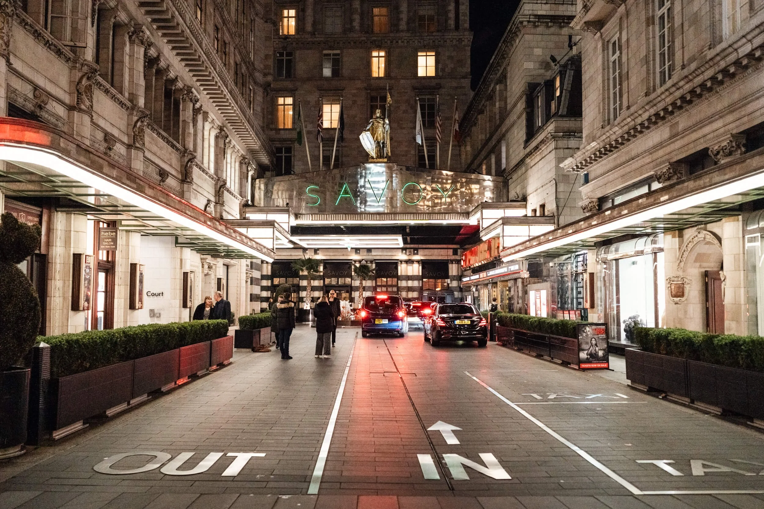 Night view of a city street entrance to a building with a green neon sign that says 'SAVOY'. Cars are parked and driving in the street, with pedestrians walking on the sidewalk.