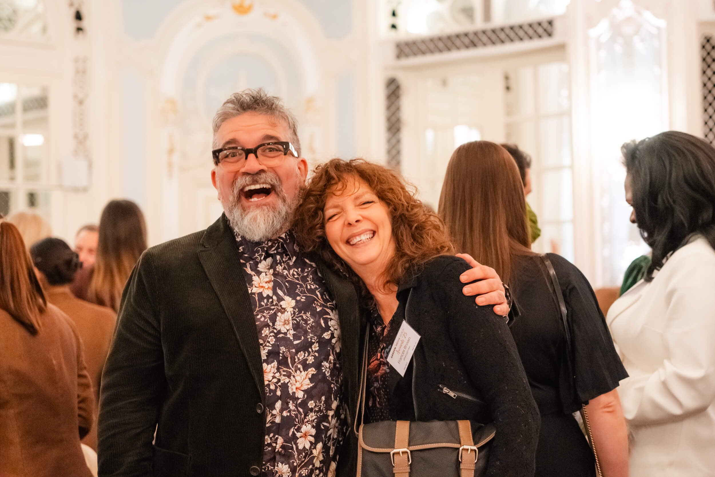 Two people smiling and hugging at a social event, surrounded by other guests, in a bright, ornate room.