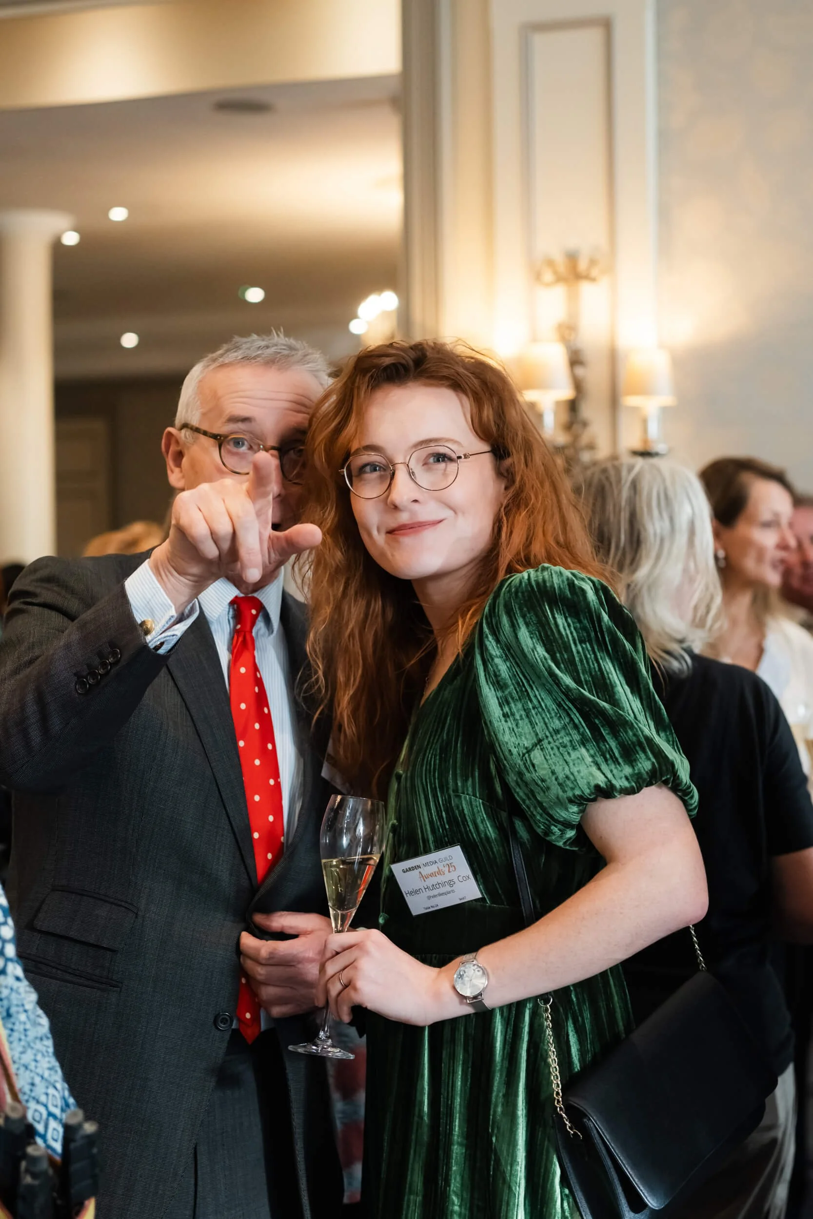 A man in a suit and a woman in a green dress posing with glasses at an event, with people in the background.