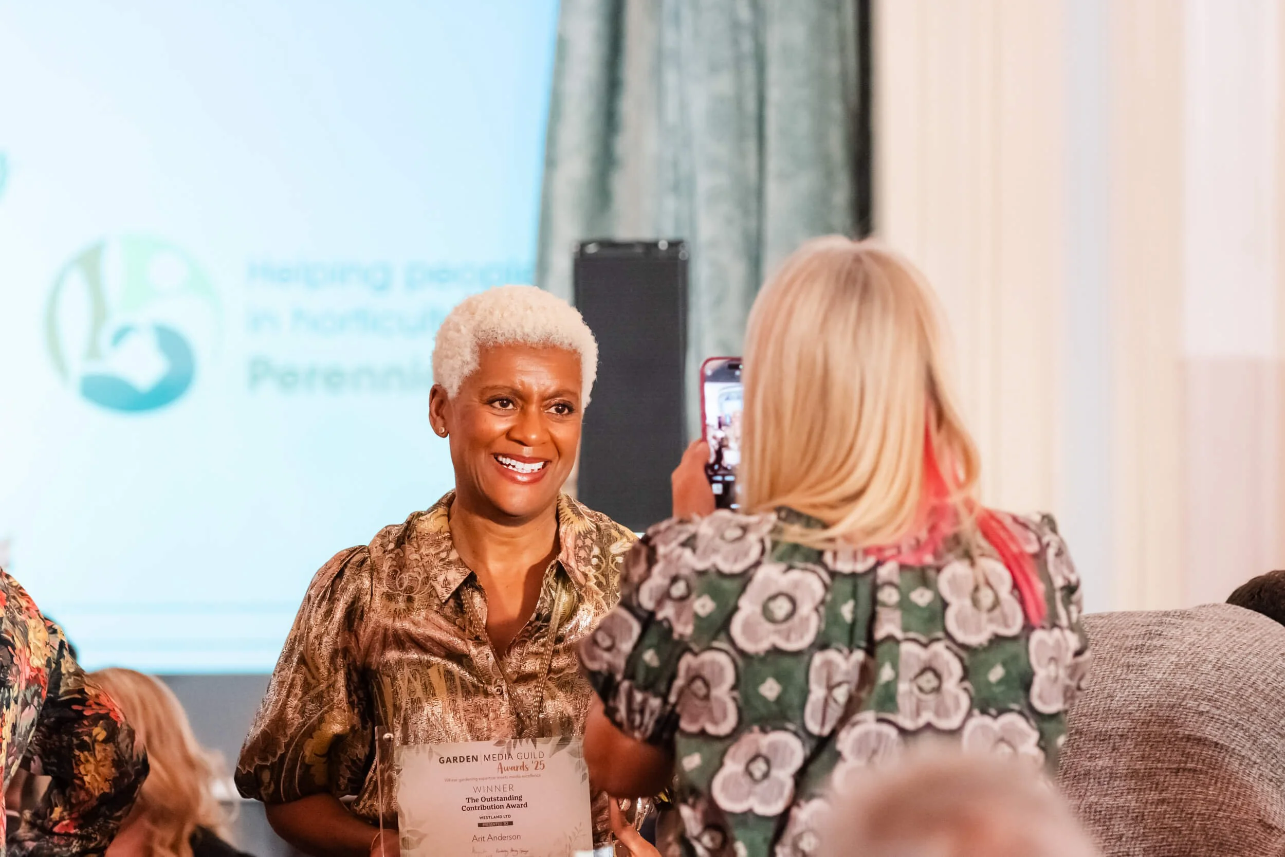 A woman with short white hair smiling as she receives an award at an event, with a woman taking a photo of her. The award has the name Art Anderson on it. In the background, a presentation screen displays the logo and text for an event related to fos