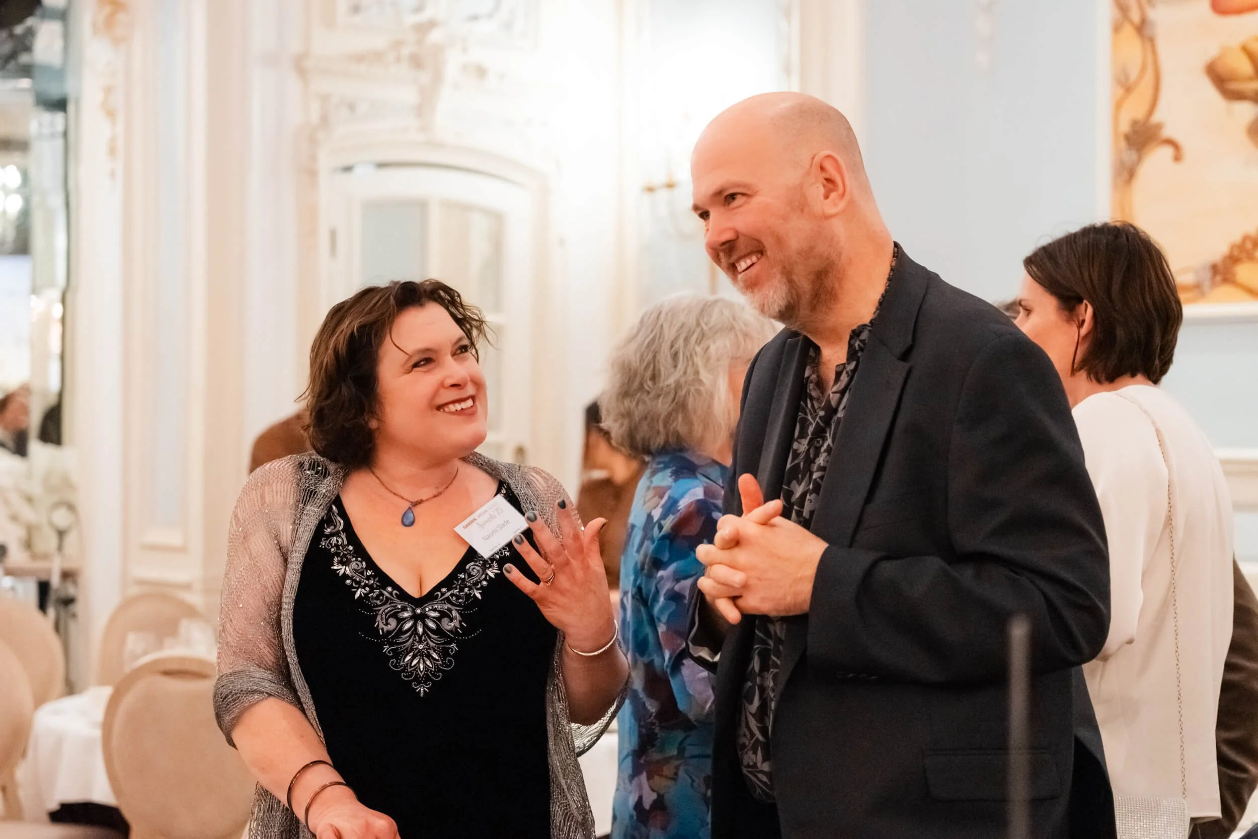 A woman and a man are engaged in conversation at a social event. The woman has curly dark hair, wears a black dress with silver embroidery, and a blue necklace. The man is bald, has a beard, and wears a black suit with a patterned shirt.