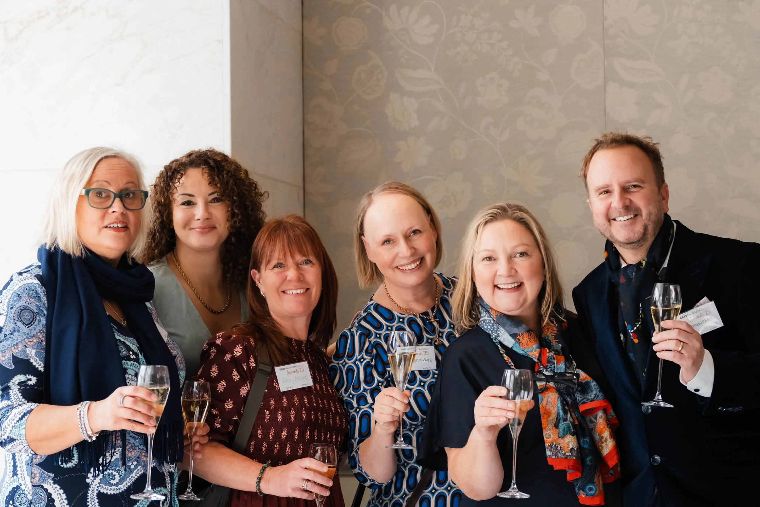 Group of seven people, six women and one man, smiling and holding glasses of champagne at a celebration or event.