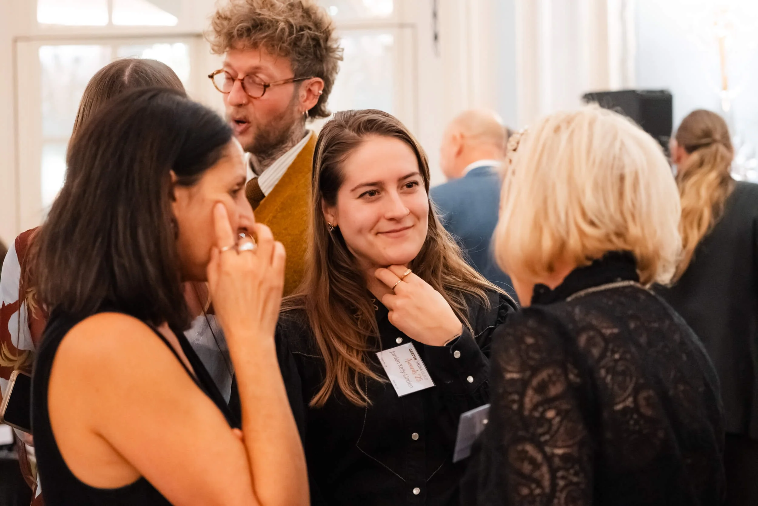 Four women and one man chatting at a social event in a well-lit room with large windows, some people in the background, and one woman with name tag.