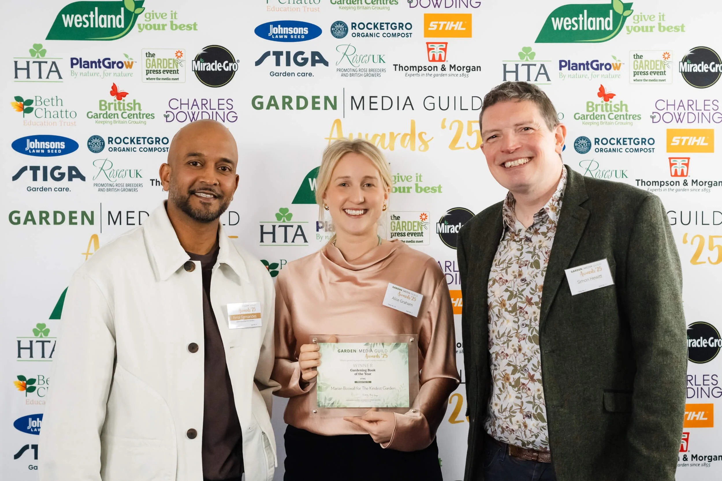 Three smiling people standing in front of a backdrop with logos, with one woman holding an award certificate. The woman in the center has blonde hair, wearing a satin pink blouse, and is holding a certificate that reads 'Garden Book of the Year.' The
