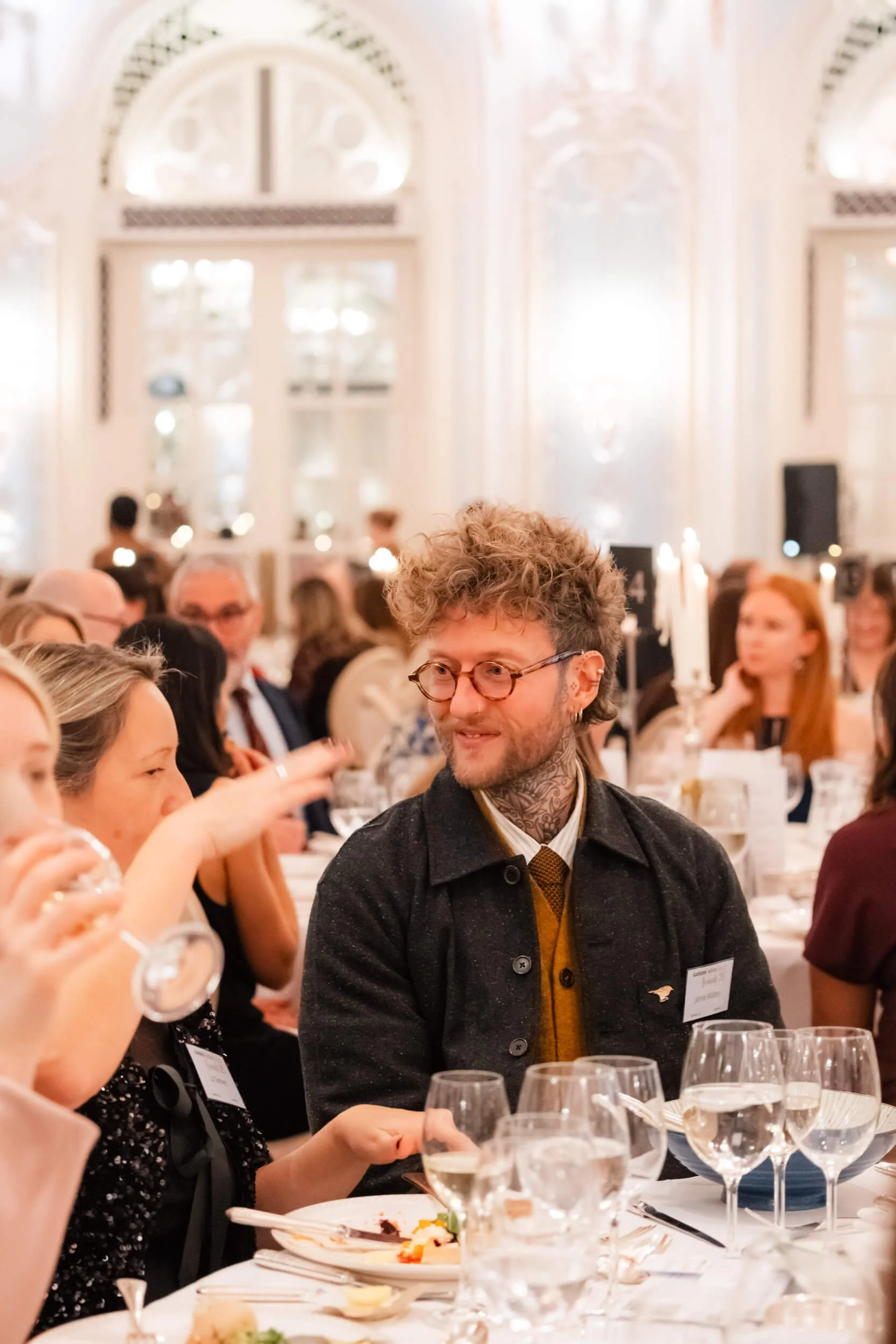 A man with curly hair, glasses, and tattoos on his neck, wearing a dark coat, is sitting at a table during a formal event, surrounded by other people, in an elegant, well-lit room with large arched windows.