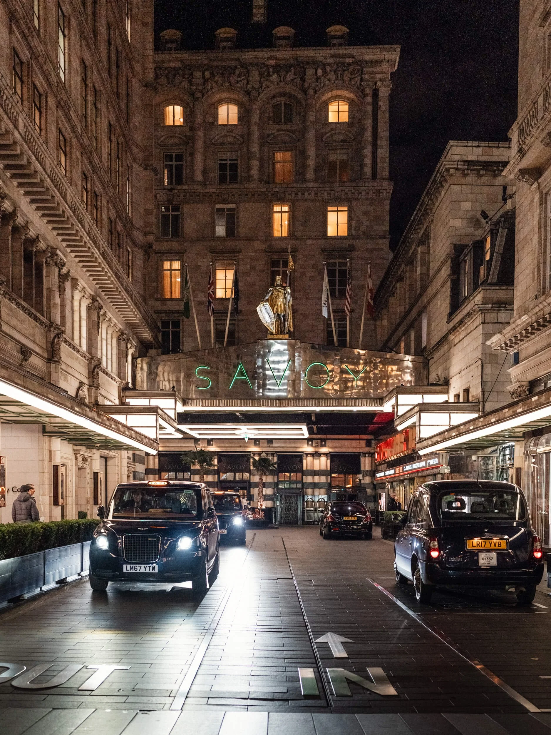 Night view of the Savoy Hotel with black taxis parked in front, illuminated hotel sign, flags, and a statue on top of the entrance