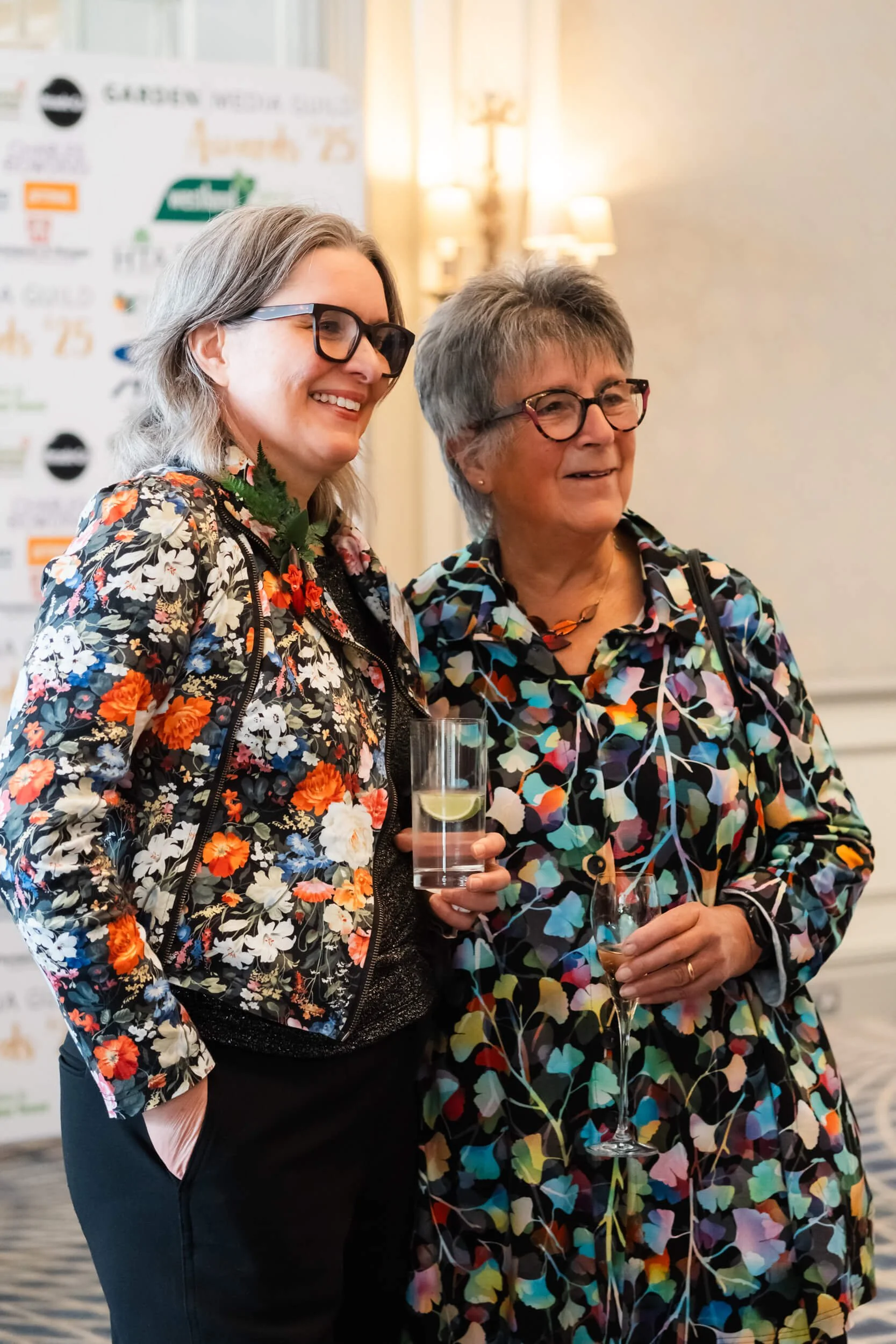 Two women wearing floral-patterned clothing stand side by side, smiling at an event. One holds a glass of water with lemon, and the other holds champagne. The background includes a banner and warm lighting.