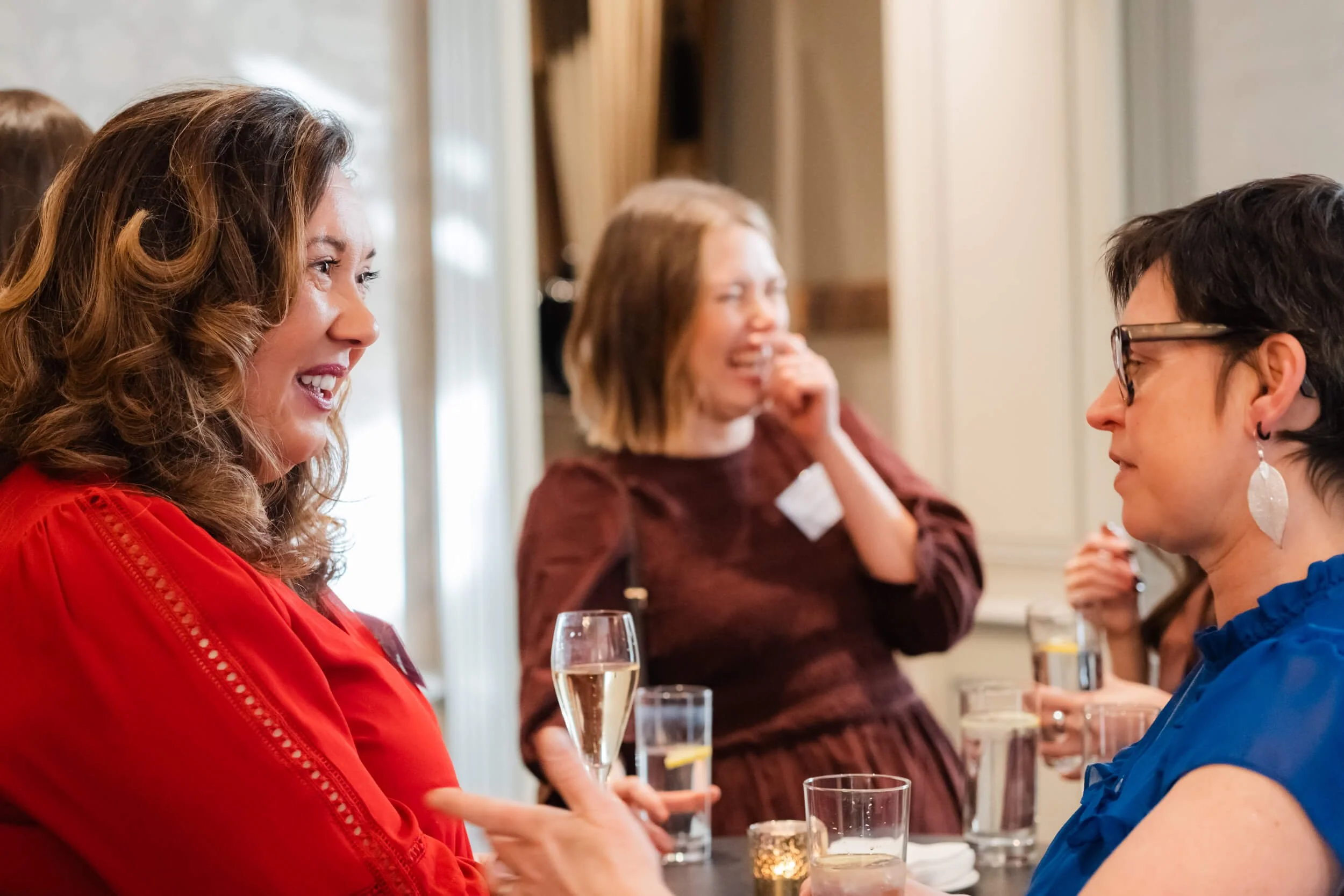 Two women are engaged in a conversation at a social gathering, smiling and holding drinks, with other women in the background also enjoying the event.